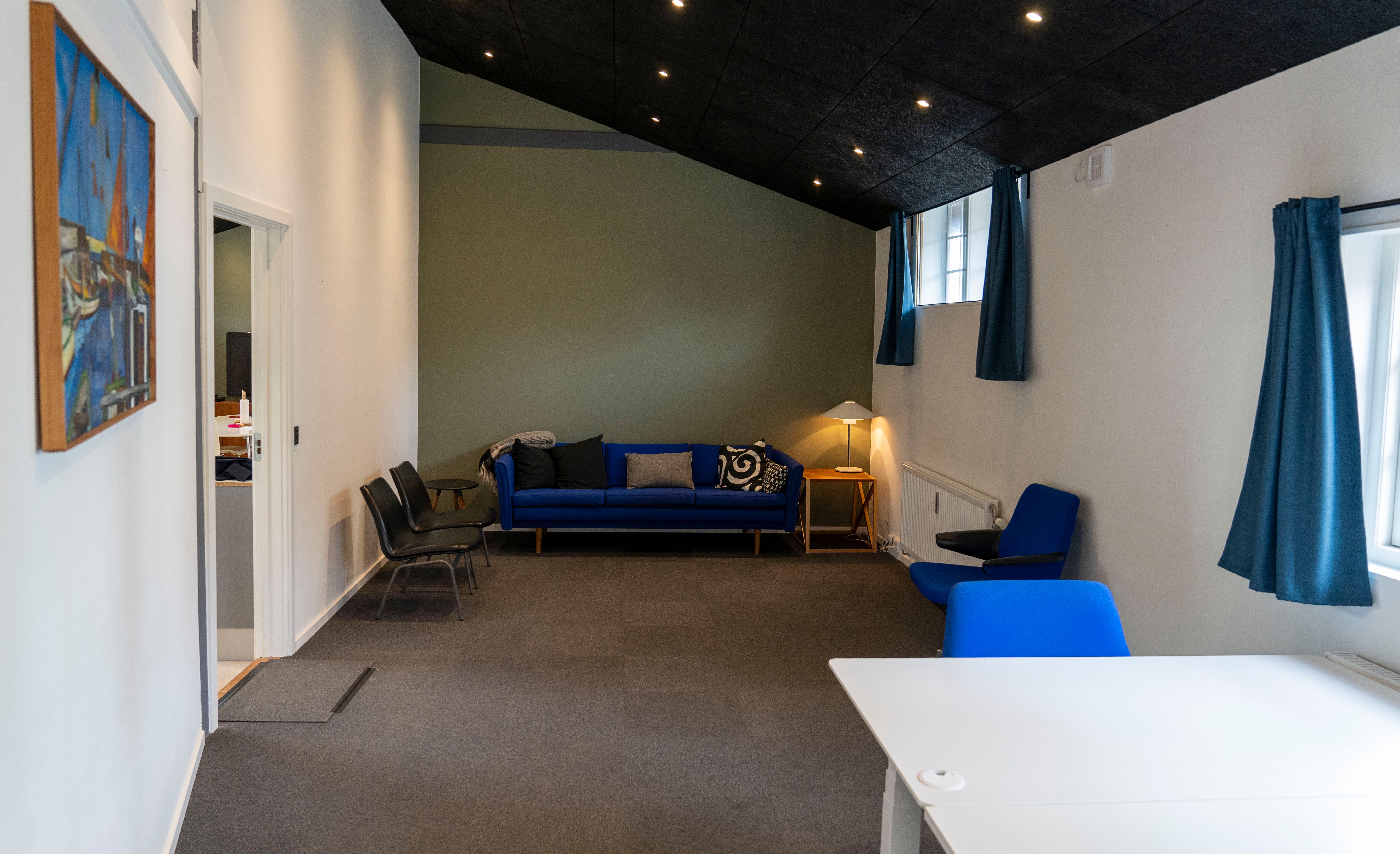 Kontorhjørne med sofa og stol. Hjørne med højt til loftet og vindue med gardiner. Billede af skibe på væggen. Væggene er