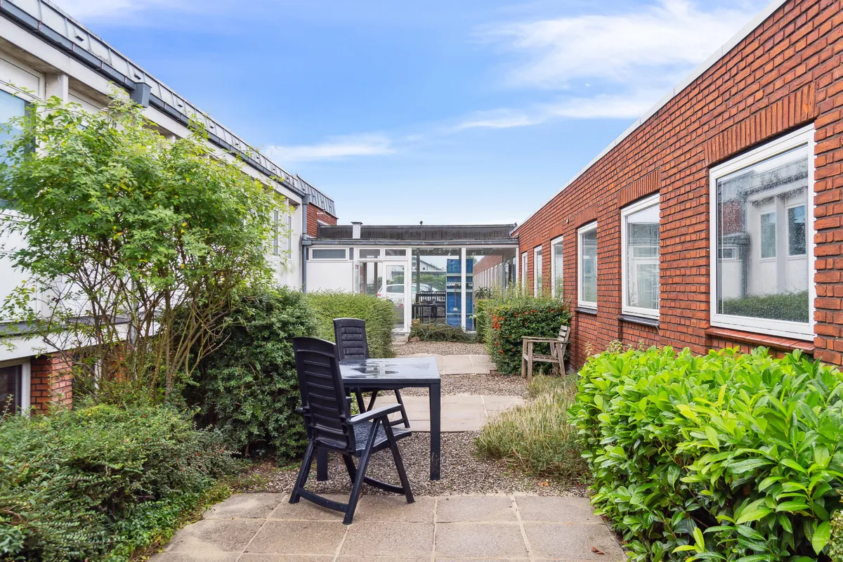 Et lille gårdhavenområde med en terrasse, udstyret med to stole og et bord, omgivet af buske og planter, ligesom der er en