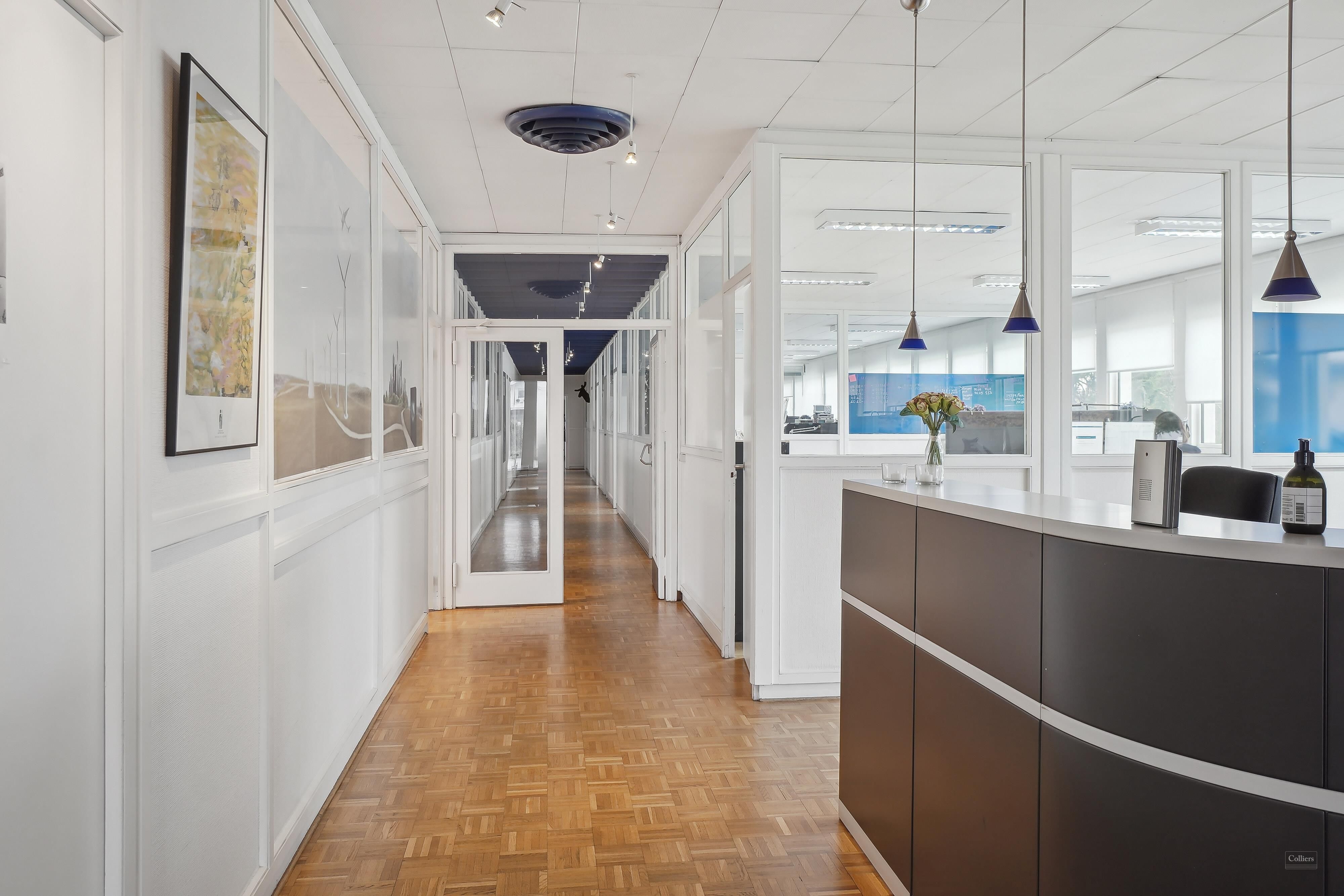 Foto af en hvidt indrettet korridor med lysekroner, malerier og et skrivebord med en vase med blomster og en flaske