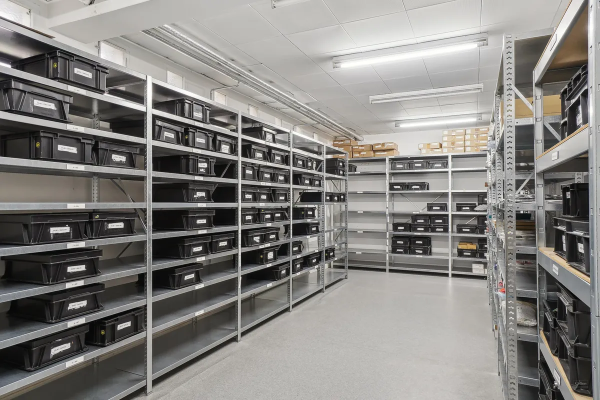 Lager med flere metalhylder, hver med sorte kasselagre, højt til loftet og godt oplyst af hvide loftslamper