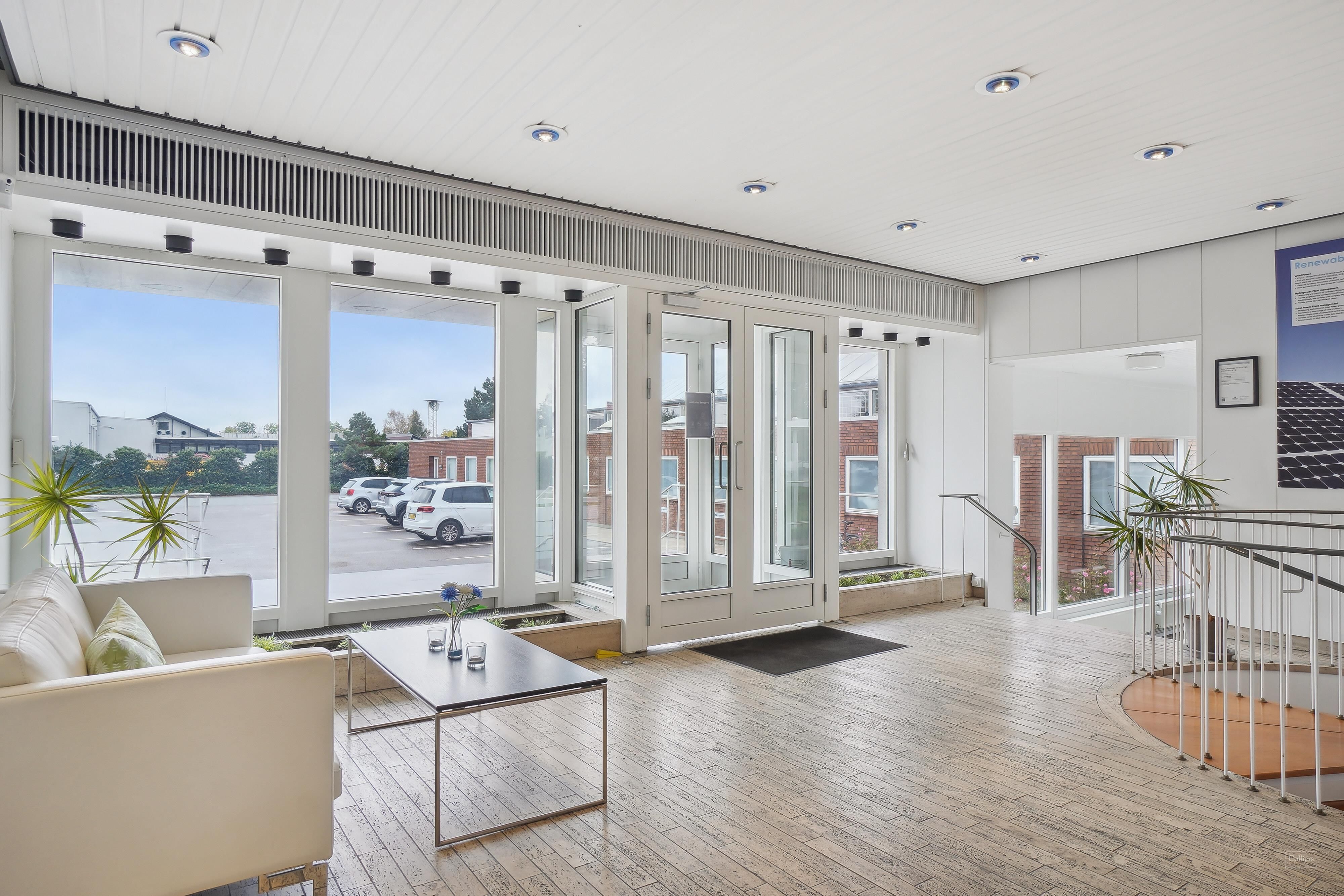 Indvendigt hjørne med hvidt sofa med pude, gulvtæppe og stol med trægulv, og vinduer med solpaneler og biler i baggrunden