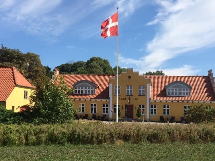 Rødt og hvidt dansk flag står på en flagpole foran en gul bygning med en rødt tag og mange vinduer