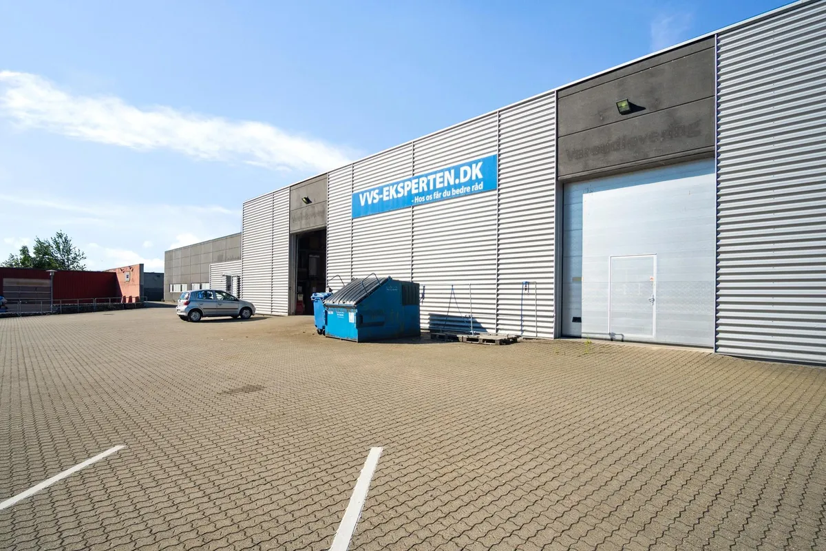 En stor lagerbygning med to store garagedøre og et bilparkeringsområde foran. Der er et skilte med teksten VVS-eksperten.dk