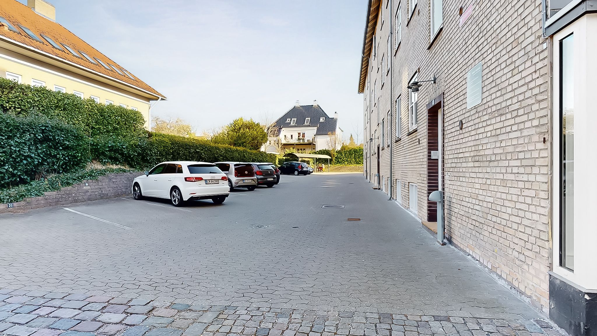 Biler parkeret på en asfaltvej mellem to bygninger med flere huse i baggrunden på en solrig dag