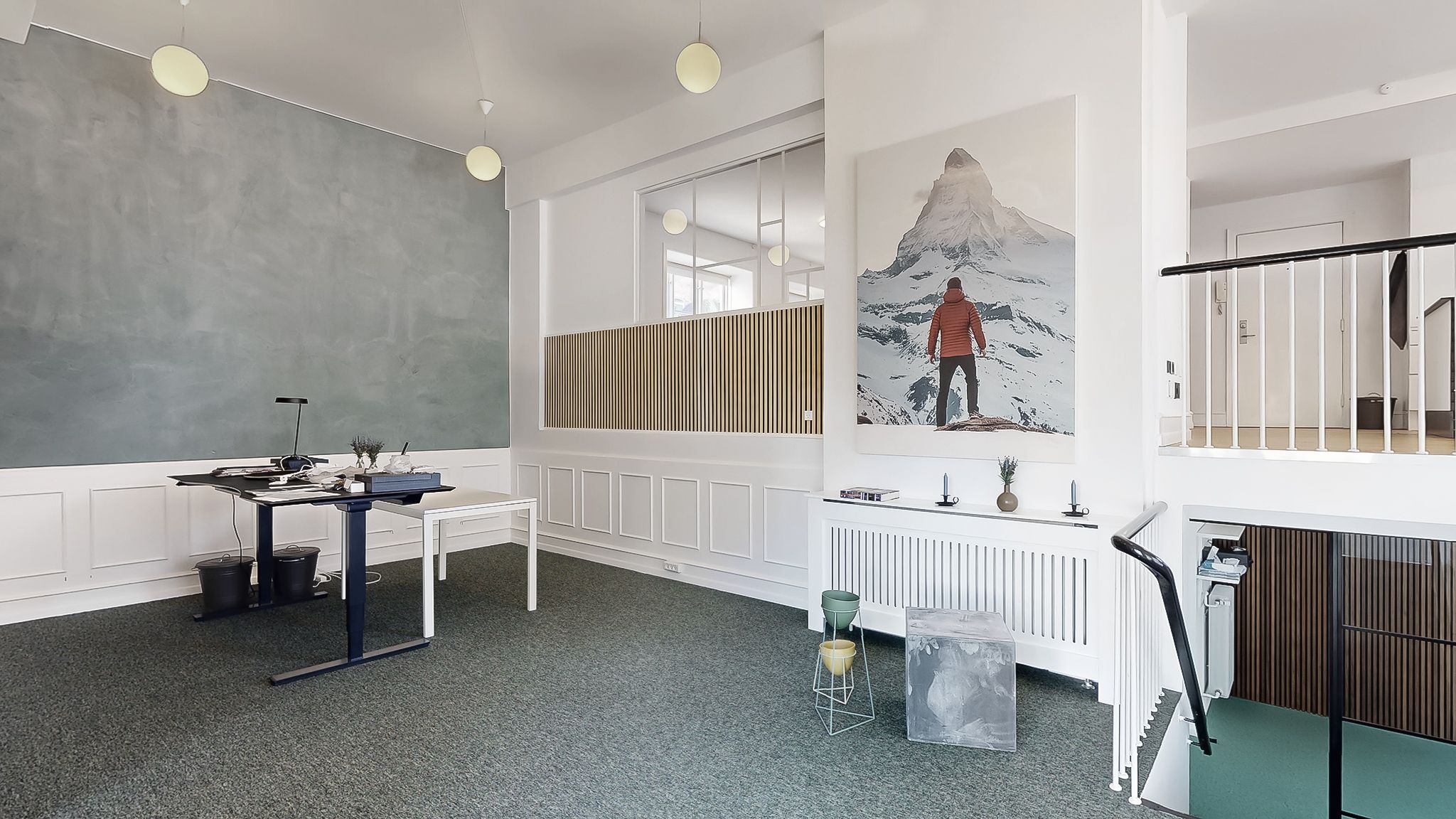 Kontorlokaler med grøn tapet, hvide vægge og et stort billede af Matterhorn på væggen. Der er en høj hylde med plante og to