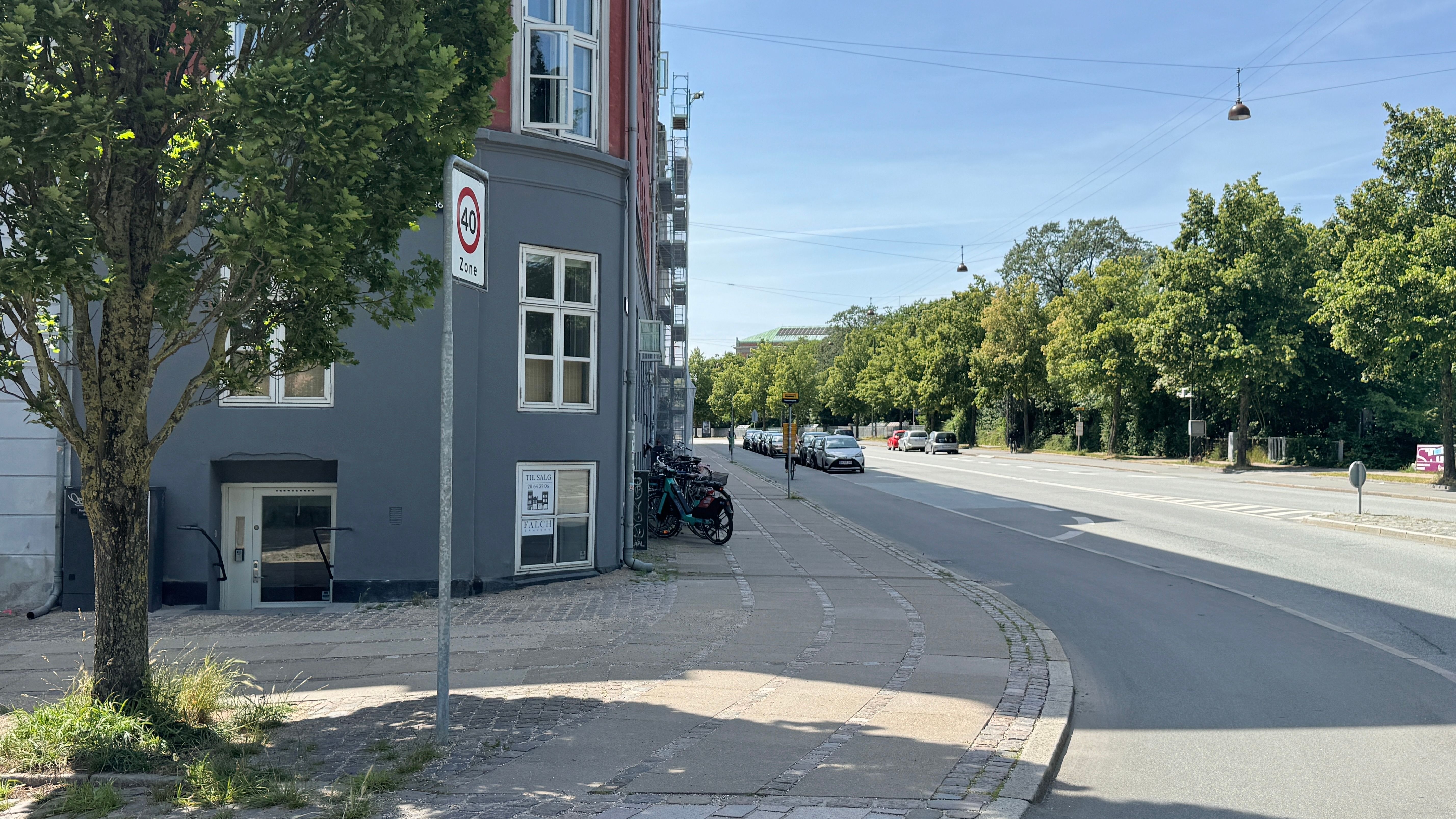 En gade med 40 km/t-grænse, parkerede biler, cykler og grønne træer, lyse himmel og sporvogne