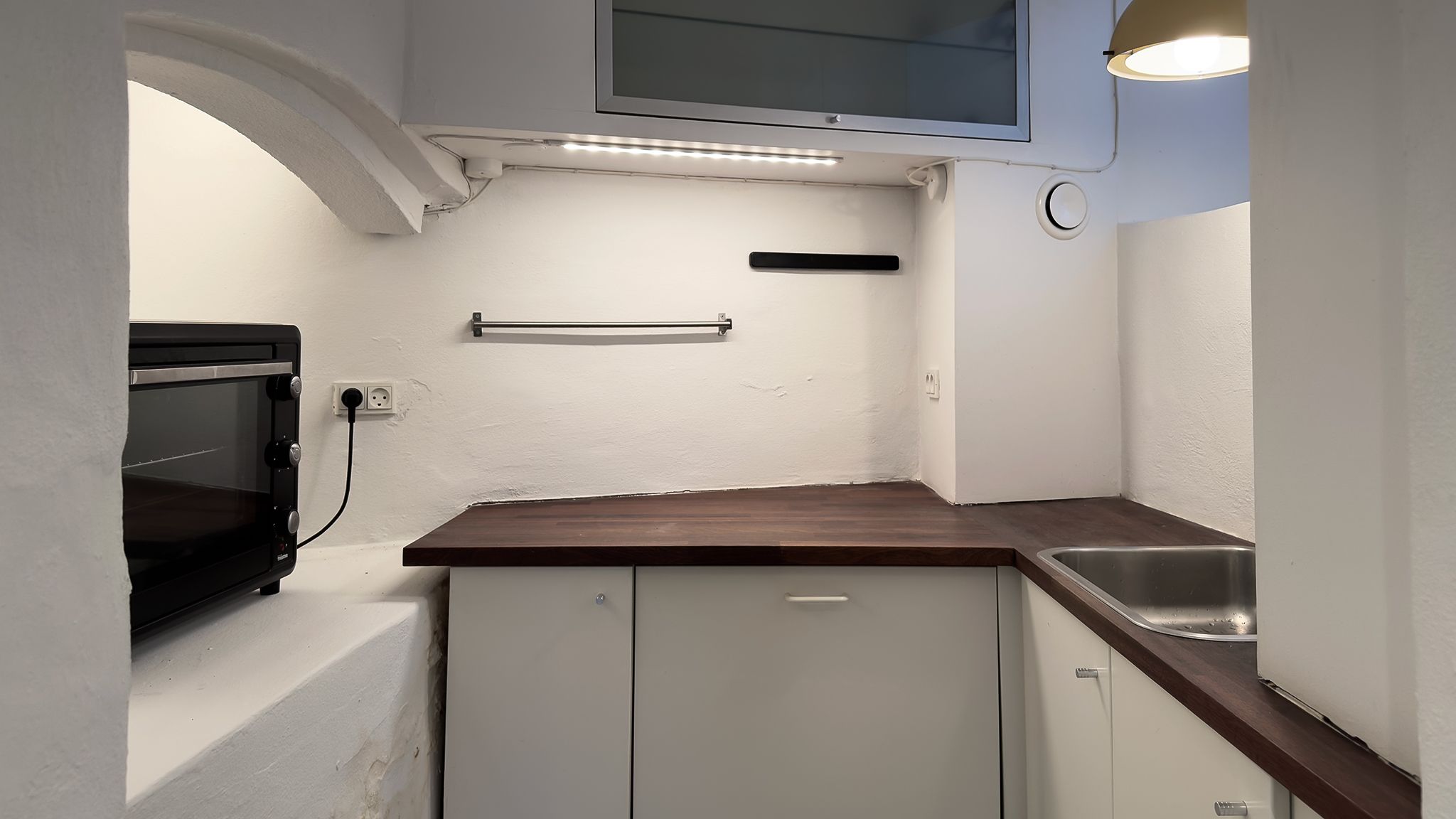 En lille køkken med hvid væg, brun træplade og lysegult loftslys, en mikroovn og et enkelt vask og underskab i hvidt