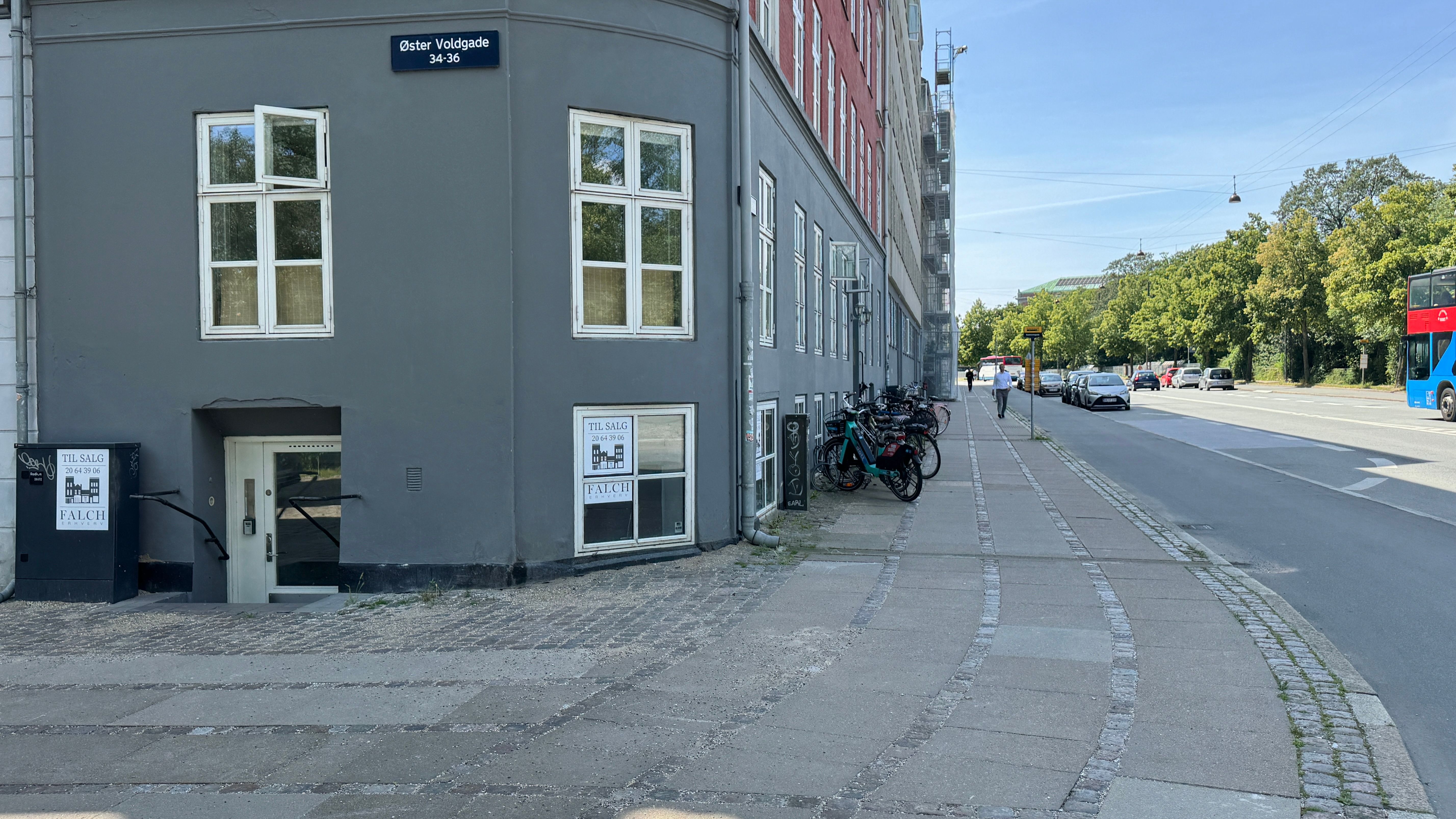 Vej og parkeringsplads med biler og en dobbeltbus ude til højre. Gågade og grønne træer. Bygning med lejlighedsnummerskilt,