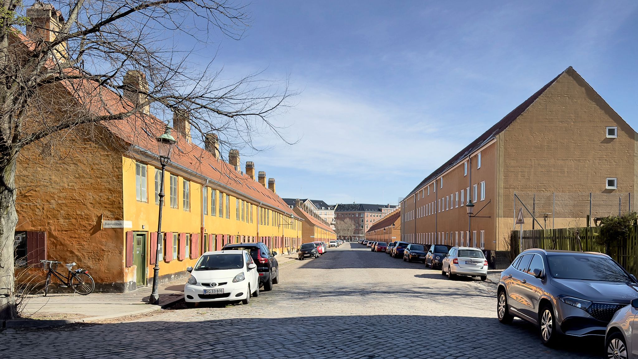 En række gule og brune husbygninger med røde ruder og røde vinduer langs Øster Voldgade, København. Parkerede biler på begge
