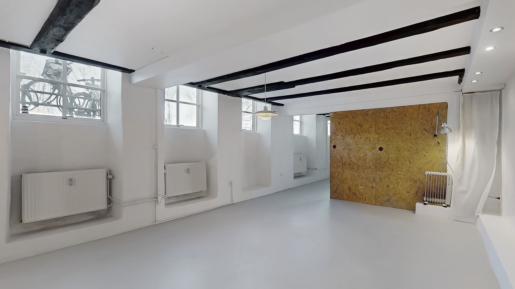 Et værelse med hvide vægge og gulv, hængende lampe og to radiatorer, et gult træpanel og en hvid gardin. To store vinduer