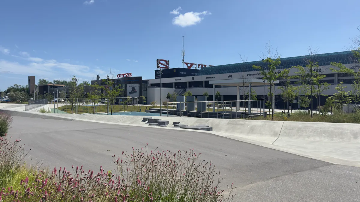Moderne bygning med City2-skilt, Intersport-skilt og H&M-skilt, samt grønne planter og blomster langs siderne