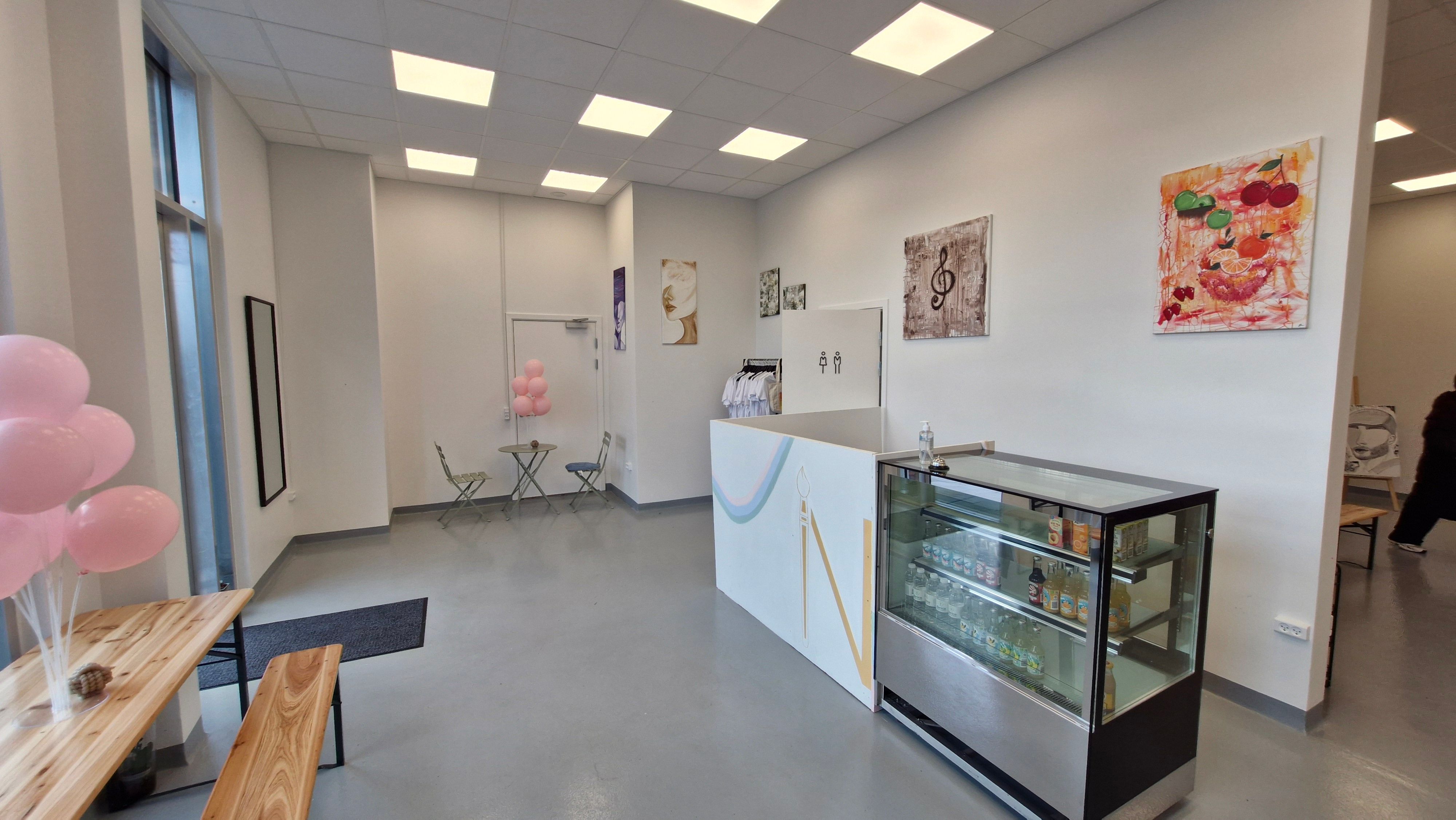 Inde i et hvidt lokale med lyse loftslamper og kunstværker på væggen. Et køleskab med drikkevarer og en bord med pink