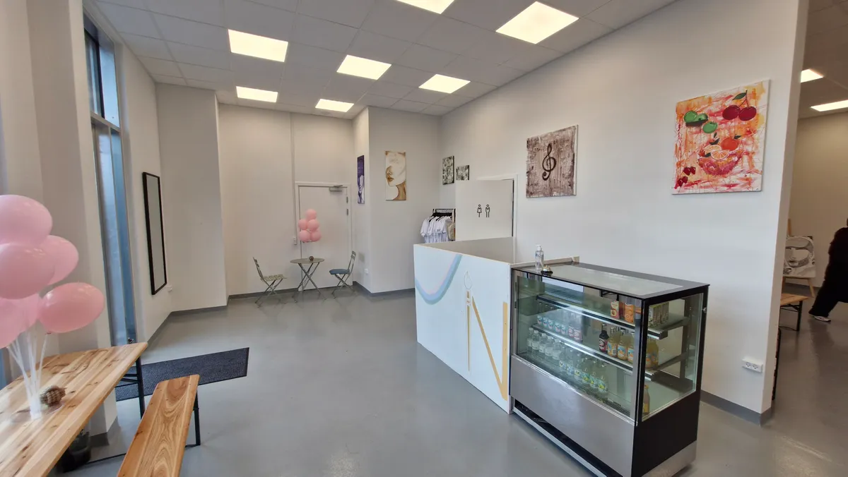 Inde i et hvidt lokale med lyse loftslamper og kunstværker på væggen. Et køleskab med drikkevarer og en bord med pink