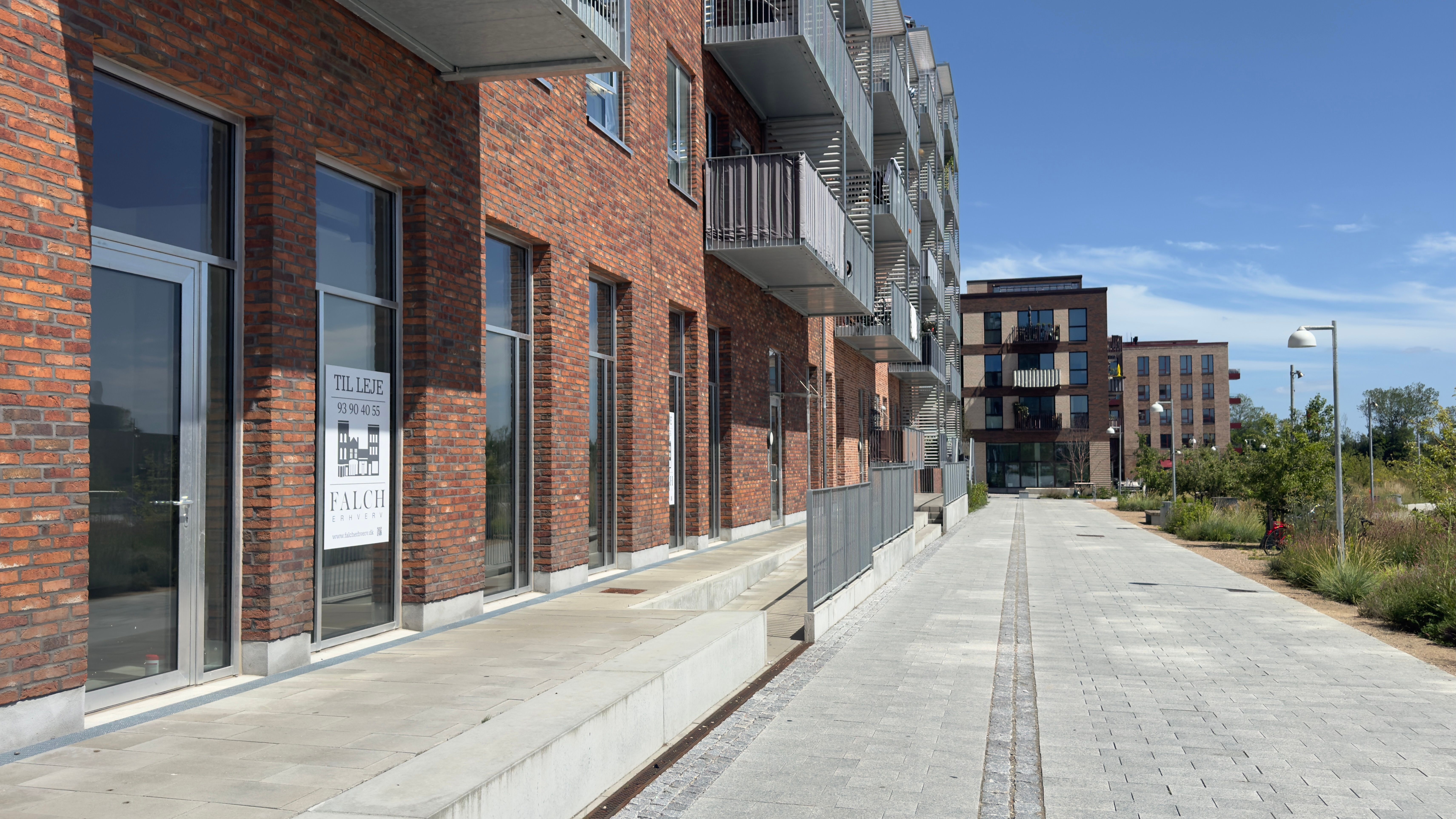 Moderne bygning med glasdøre og balkoner, sammen med en brolægget gågade og vejsideplantninger