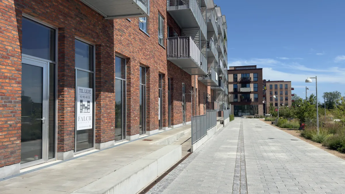 Moderne bygning med glasdøre og balkoner, sammen med en brolægget gågade og vejsideplantninger