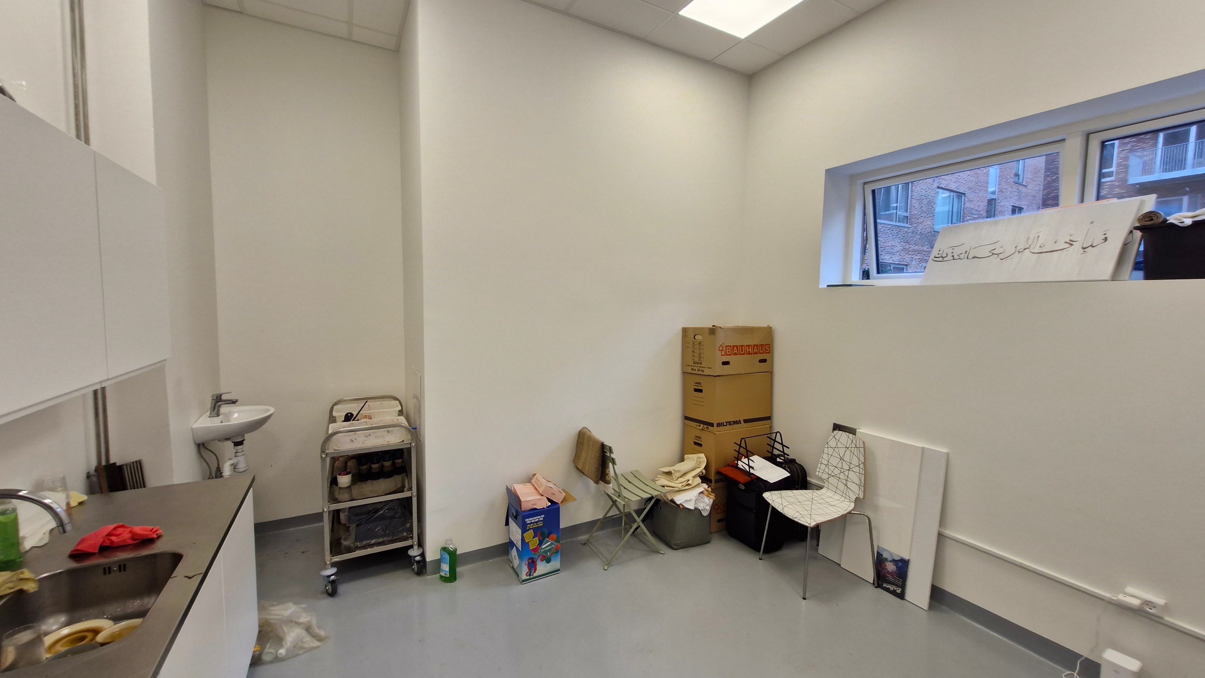 Et tomt rum med hvide vægge, vinduer, skraldespand, toilet, håndvask, køkkenbord, stålampe, hængekøkken og flere bokse og