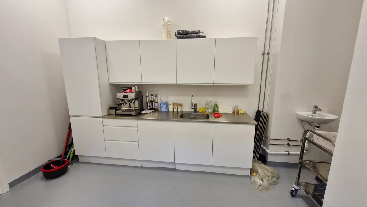 Moderne køkken med hvide vægge, hvide skuffer, lysegrå gulv og ren udsigt. Der er en kaffemaskine, en vandhane og et vask på