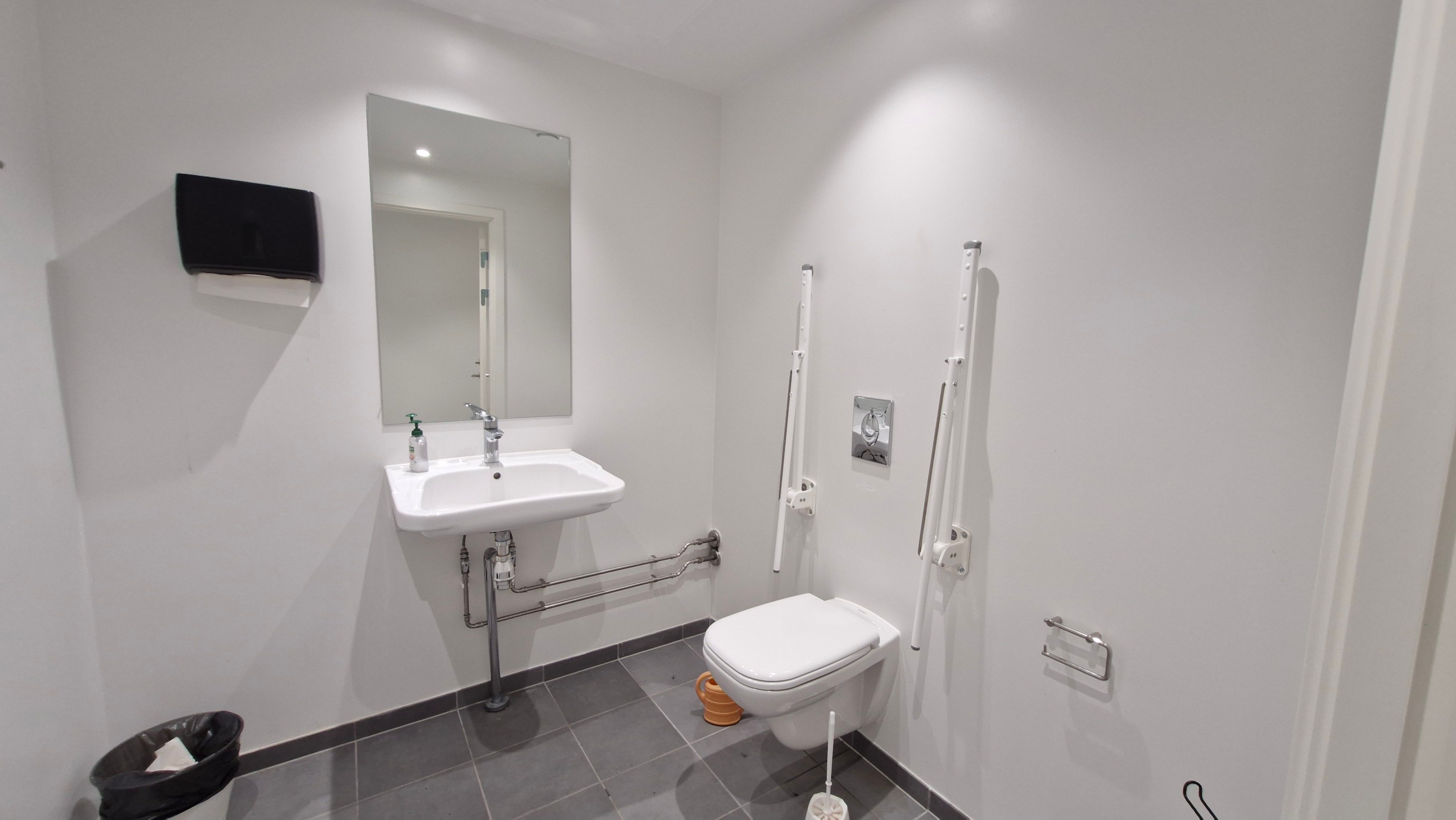 Moderne hvidt handicapvenligt toilet med håndklædeholder, håndvask, toiletbørste og to toilethåndtag, samt sæbeflaske og