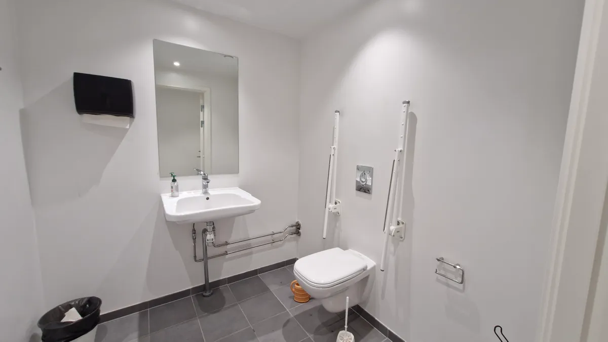 Moderne hvidt handicapvenligt toilet med håndklædeholder, håndvask, toiletbørste og to toilethåndtag, samt sæbeflaske og