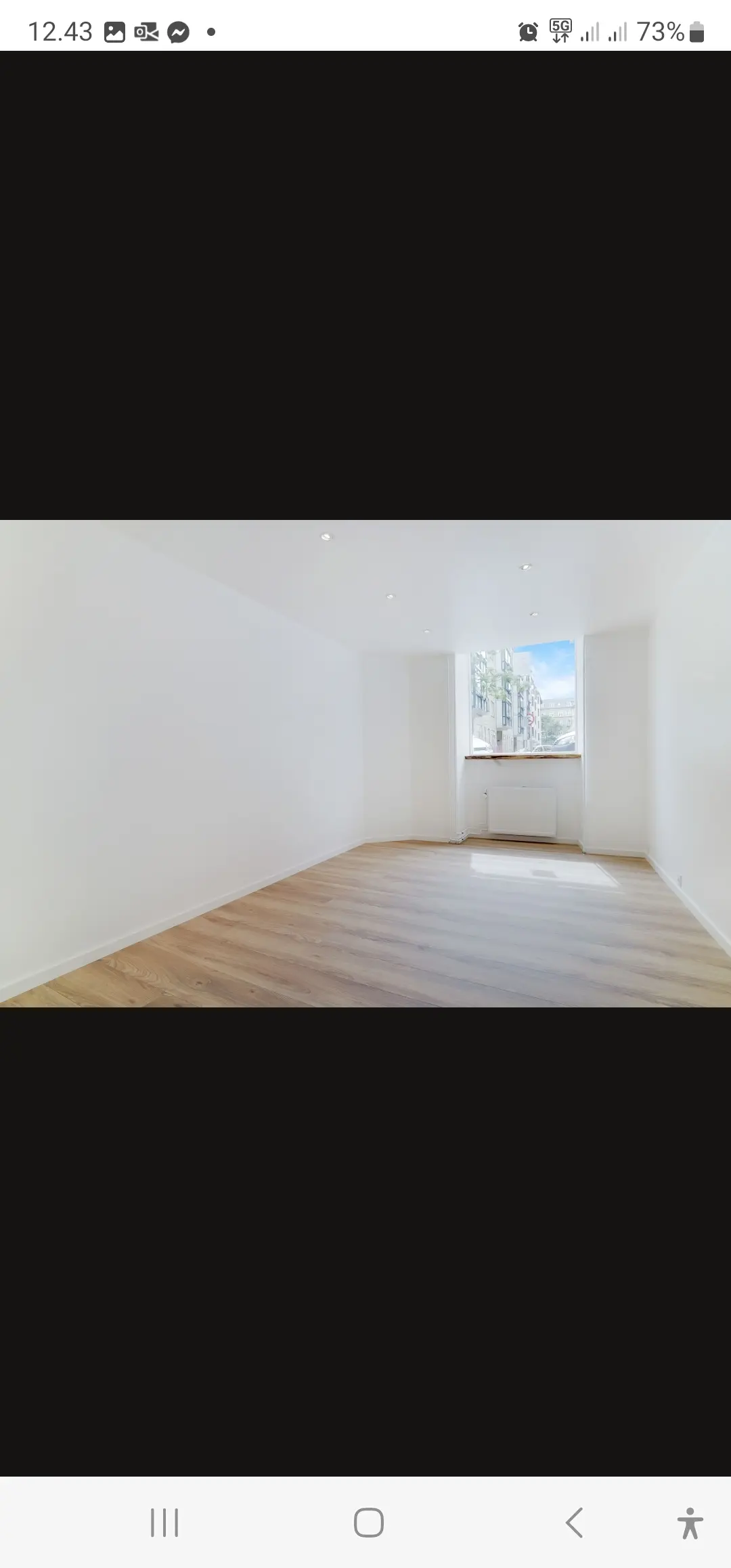 en tom stue med lysegult gulv og hvide vægge samt et vindue med udsigt til bygninger udenfor