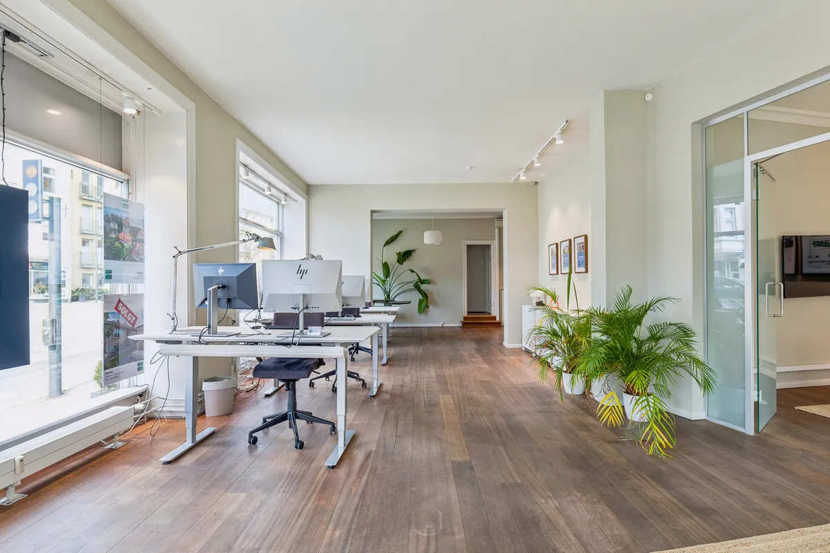 Et slankt kontor med høje ruder og lyse lofter, hvor der er to høje skriveborde med computere og stole. Der er plantegninger