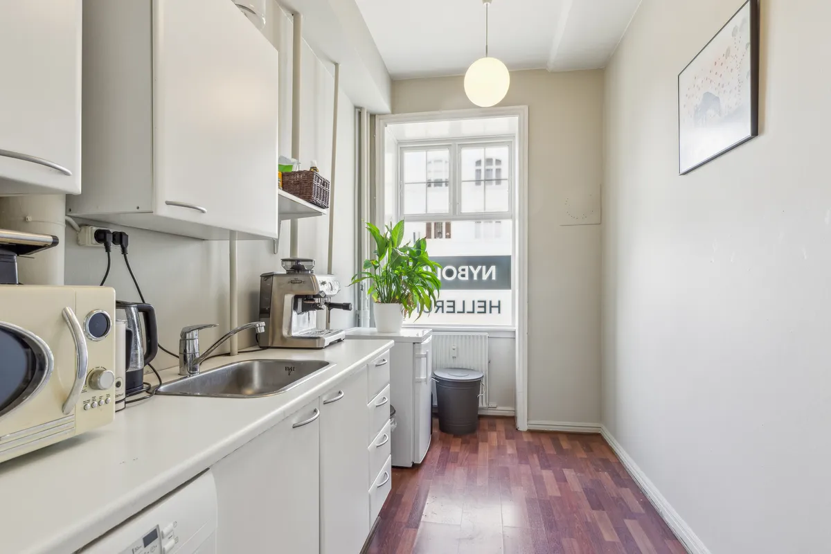 Et lille, moderne køkken med hvide skabe og lysegul gulvbelægning, hvor der er en mikrobølgeovn og en kaffemaskine på disken