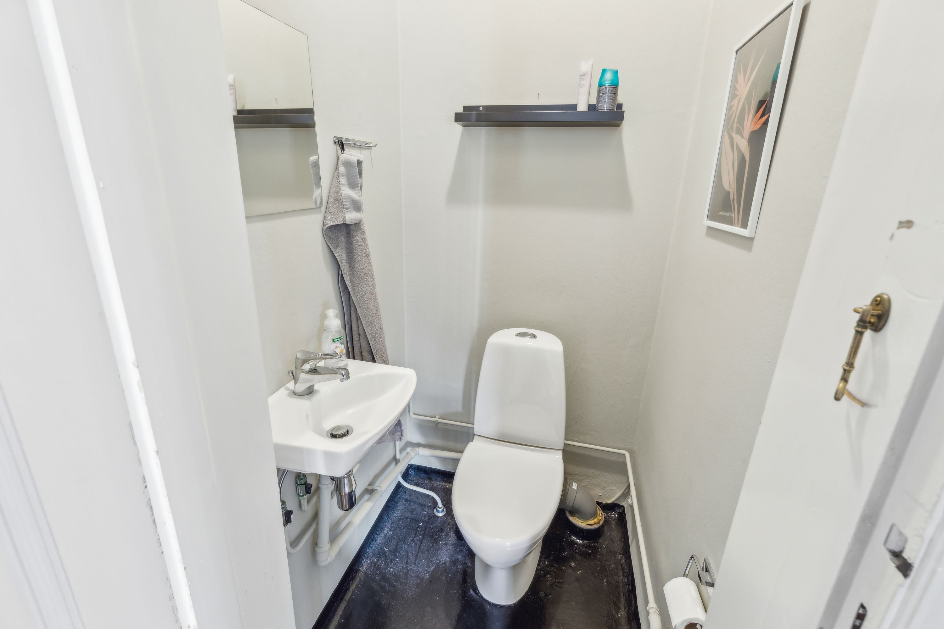 Et lille badeværelse med en hvid toilet, en hvid vask, et spejl og en håndklædeholder. Et billedramme hænger på væggen og en