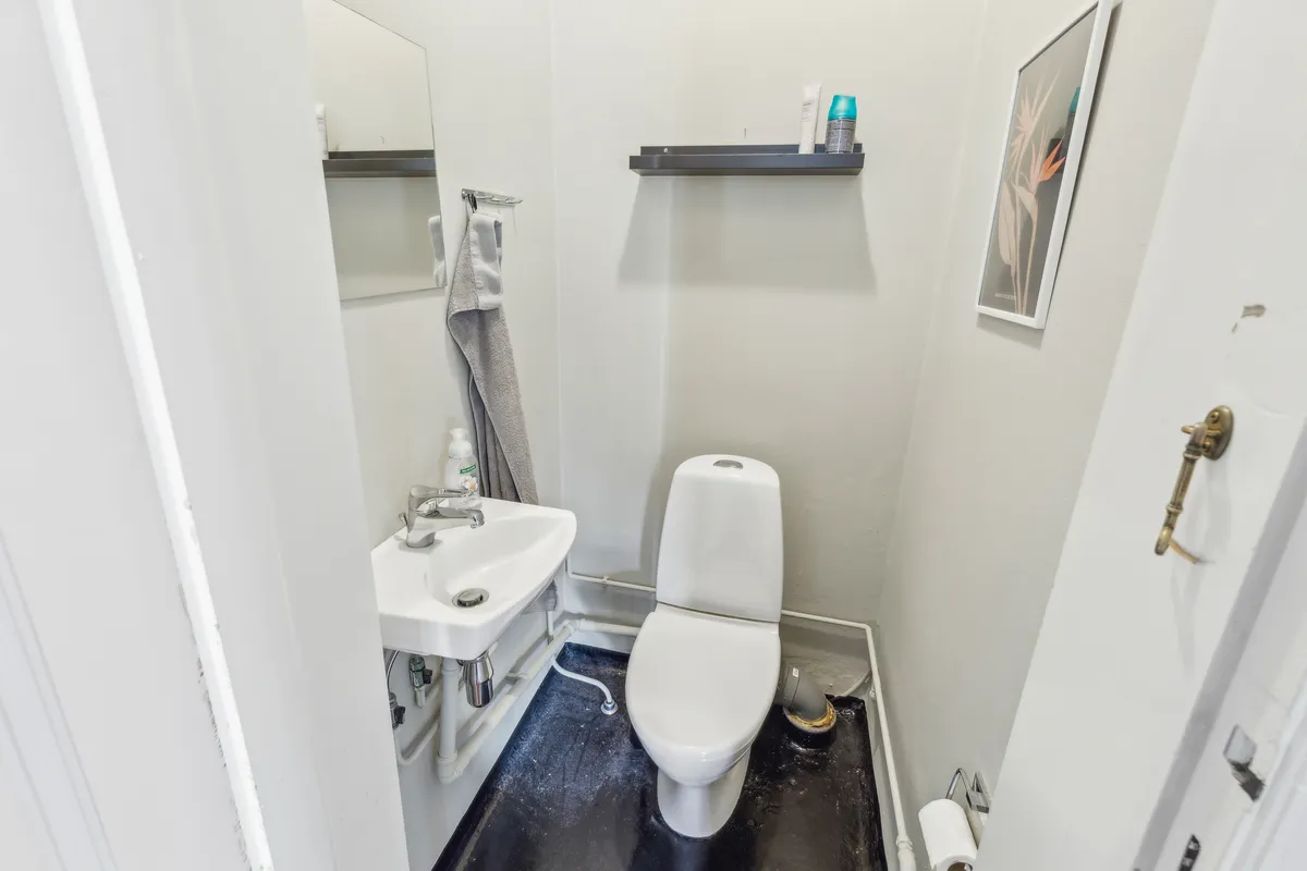 Et lille badeværelse med en hvid toilet, en hvid vask, et spejl og en håndklædeholder. Et billedramme hænger på væggen og en