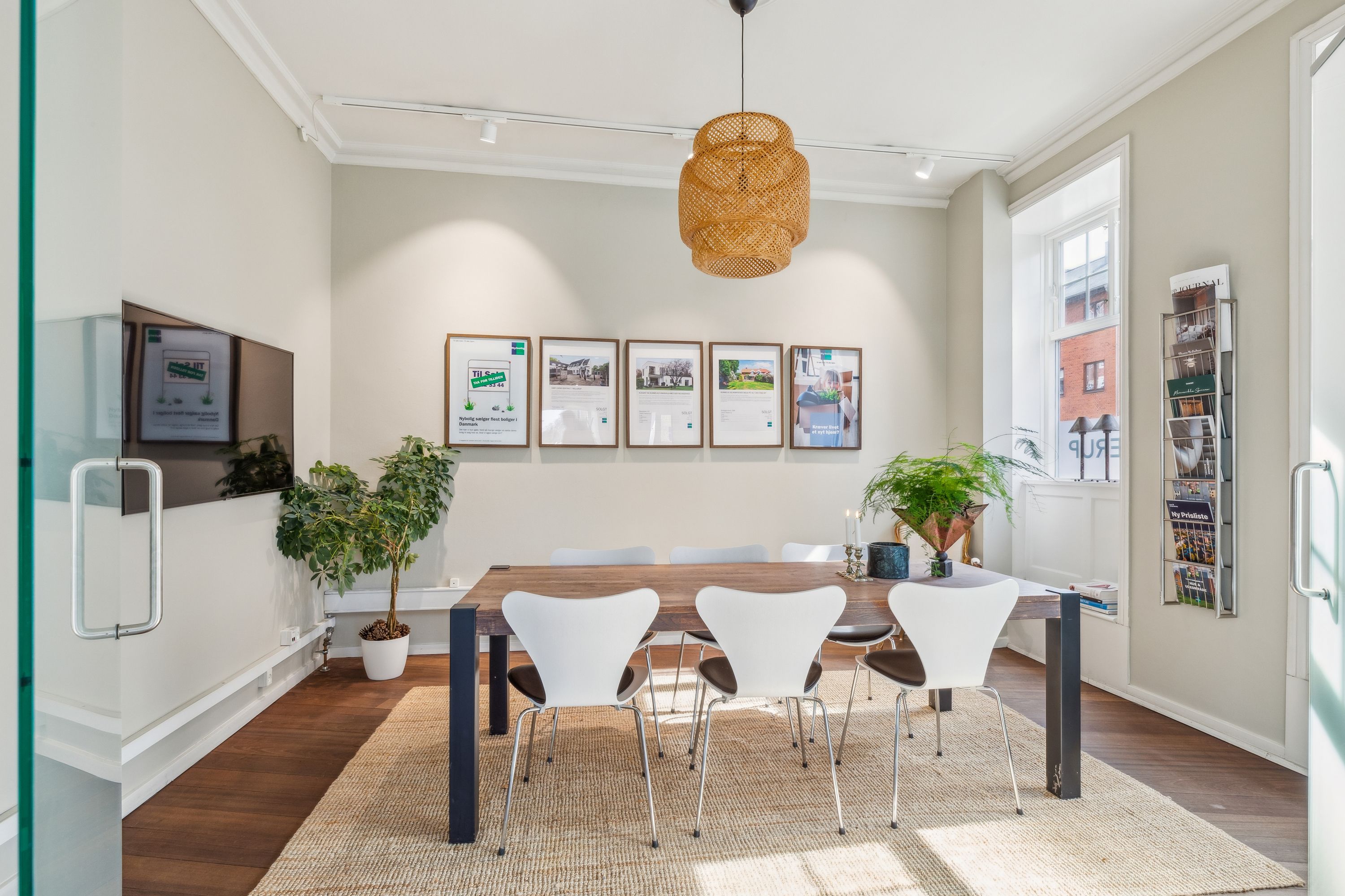 Et moderne kontorlokal med lyse vægge og hårdt trægulv. Værelset har et rundt træbord med hvide stole og en lysekron. Der er