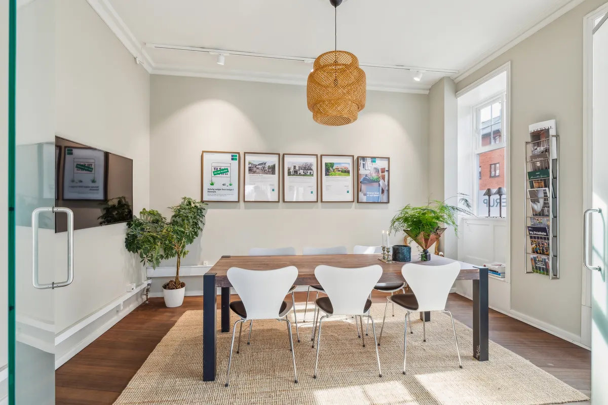 Et moderne kontorlokal med lyse vægge og hårdt trægulv. Værelset har et rundt træbord med hvide stole og en lysekron. Der er