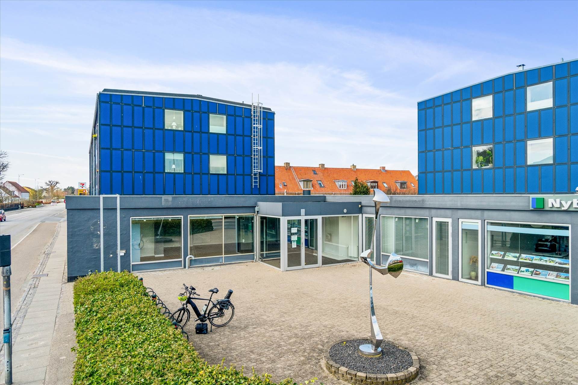 To cykler parkeret foran et bygningskompleks med en hjørnebygning i blåt og en gul facade med et kunstværk foran