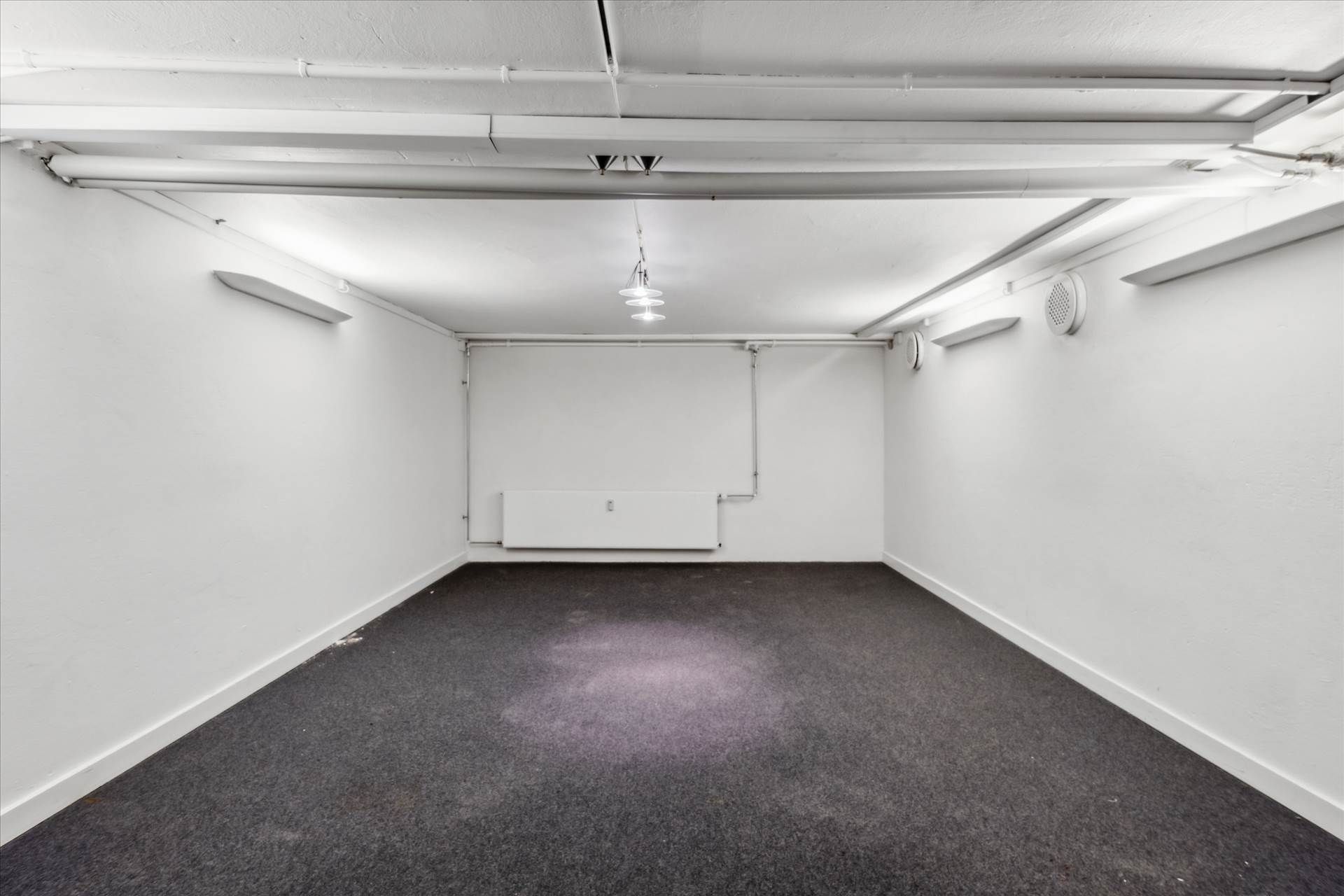 Et tomt rum med hvide vægge og højloft. Gulvet er dækket med lysegråt teppe og lysekroner hænger fra loftet. Der er hvide