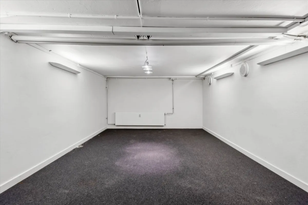 Et tomt rum med hvide vægge og højloft. Gulvet er dækket med lysegråt teppe og lysekroner hænger fra loftet. Der er hvide