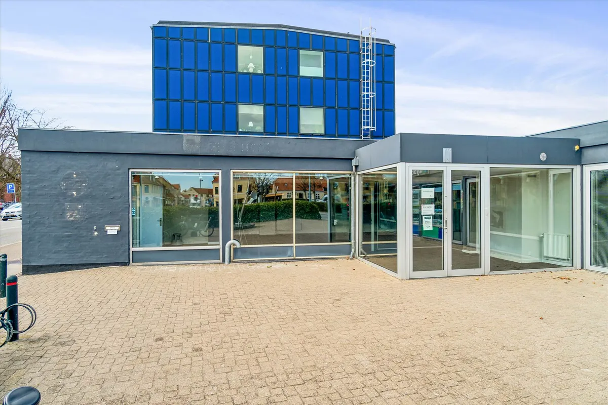Glasfasade med åbne døre, gulv med gul puds, høj blå bygning med mange vinduer og en trappe til loftet