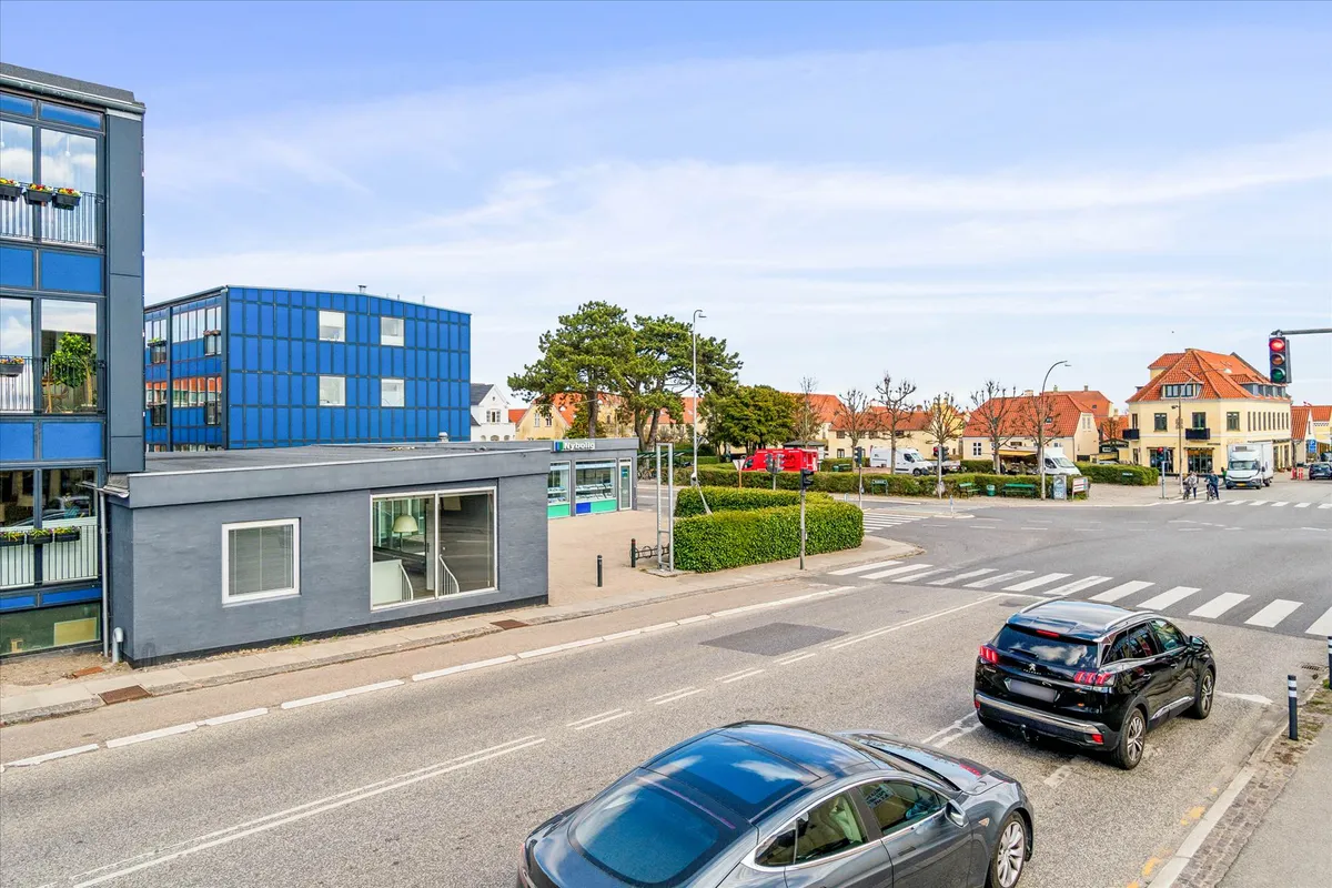 To biler kører på krydset i en by med røde taghuse og en stor blå bygning