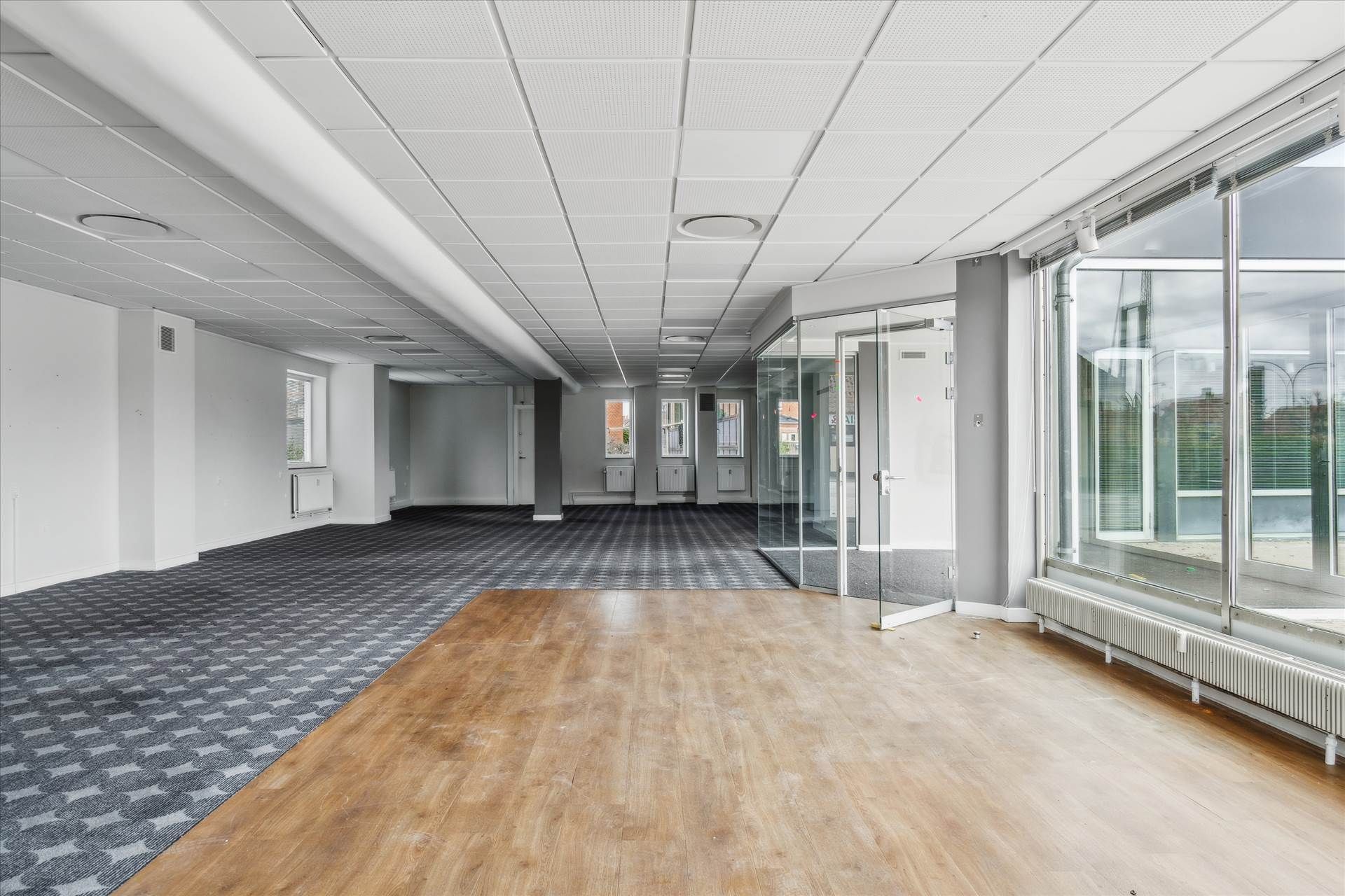 Et stort tomt rum med glasvinduer, hvide vægge og trægulve. Der er store møbler i rummet, og det er fuldstændig tomt.