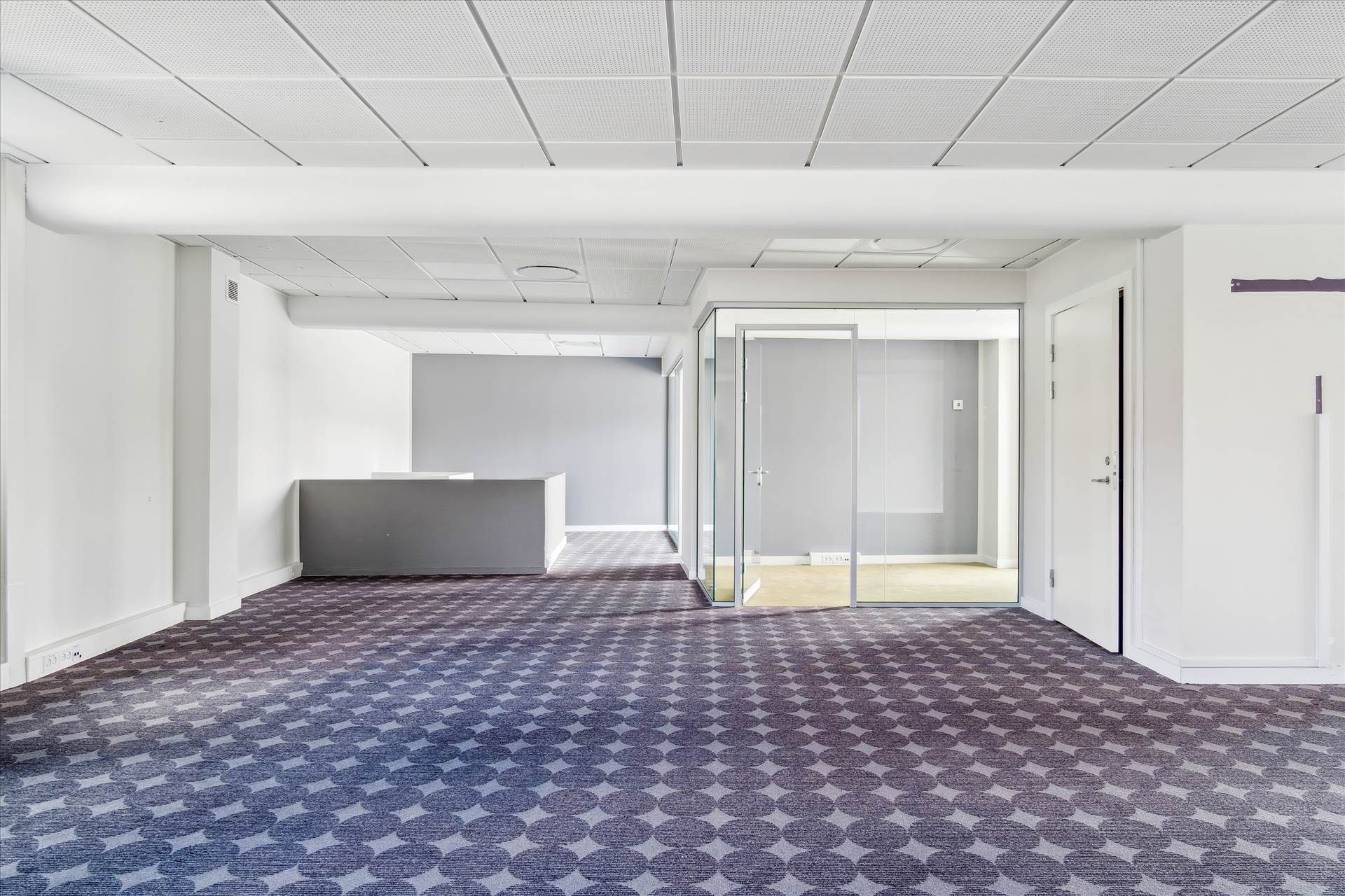 Tomt rum med gulvbelægning og glasdøre, hvidt loft, mørke vægge og et skrivebord med høj. En slank gulvbelægning med