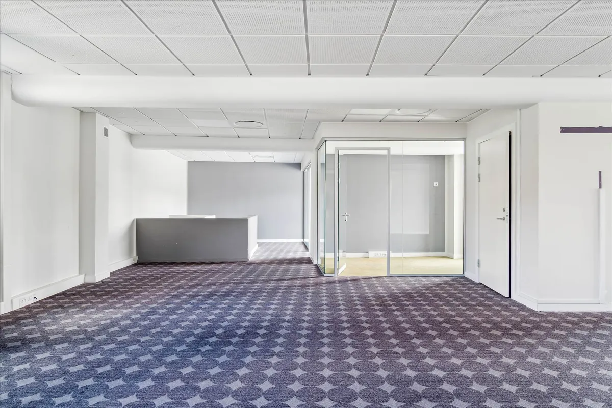 Tomt rum med gulvbelægning og glasdøre, hvidt loft, mørke vægge og et skrivebord med høj. En slank gulvbelægning med