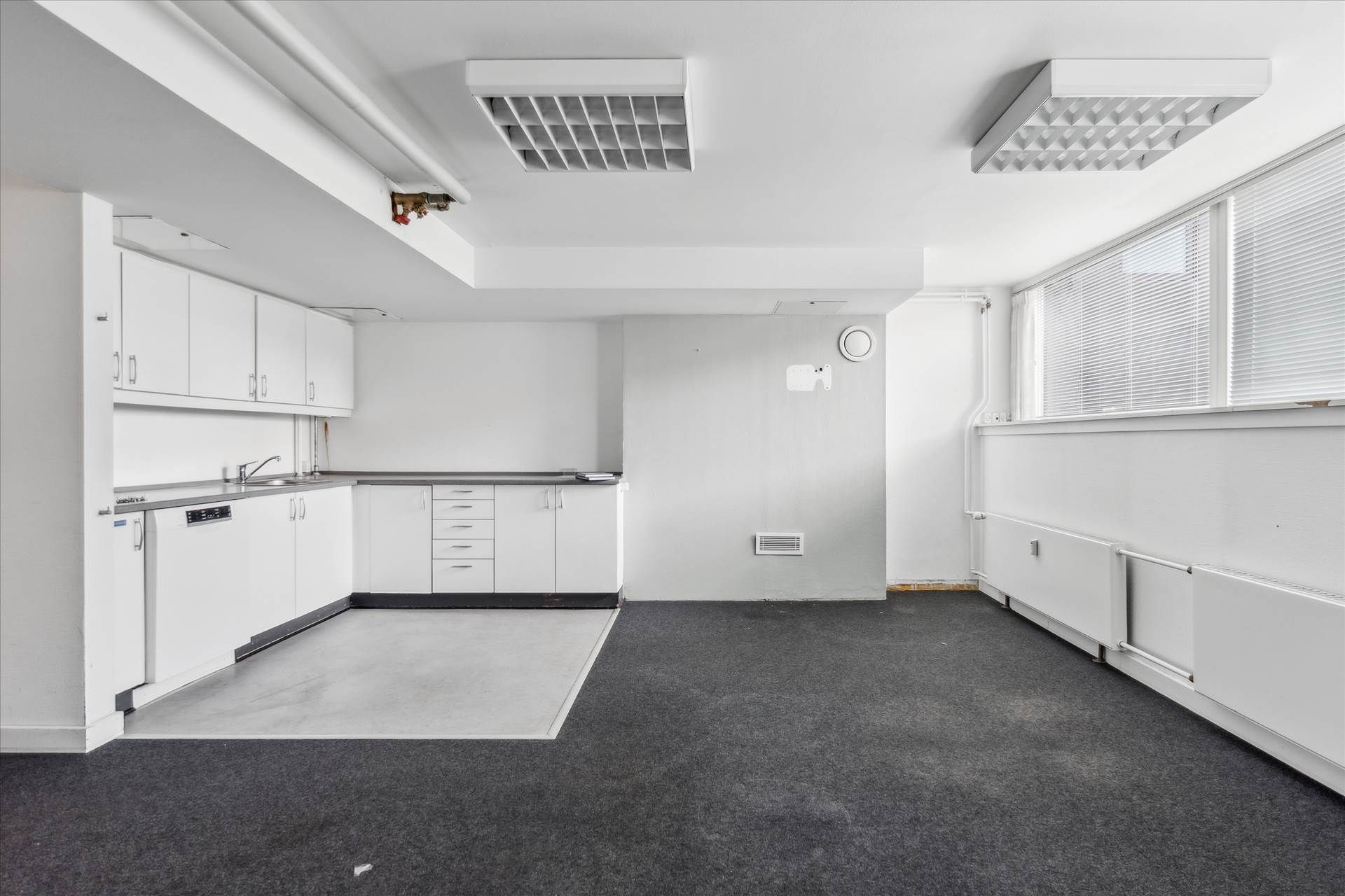 En moderne køkkenafdeling med hvide vægge, hvide skabe og en lysegrå gulv. Et hjørne med to vinduerne og en hvid radiator.