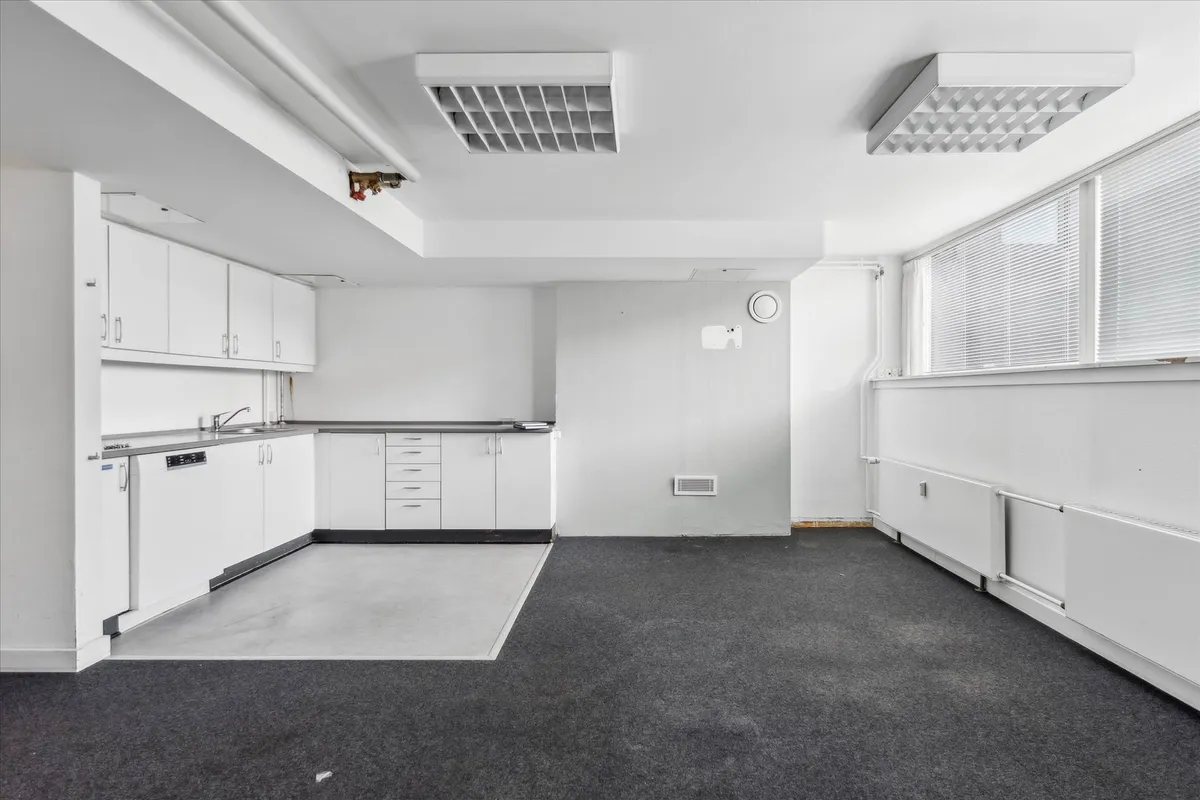 En moderne køkkenafdeling med hvide vægge, hvide skabe og en lysegrå gulv. Et hjørne med to vinduerne og en hvid radiator.