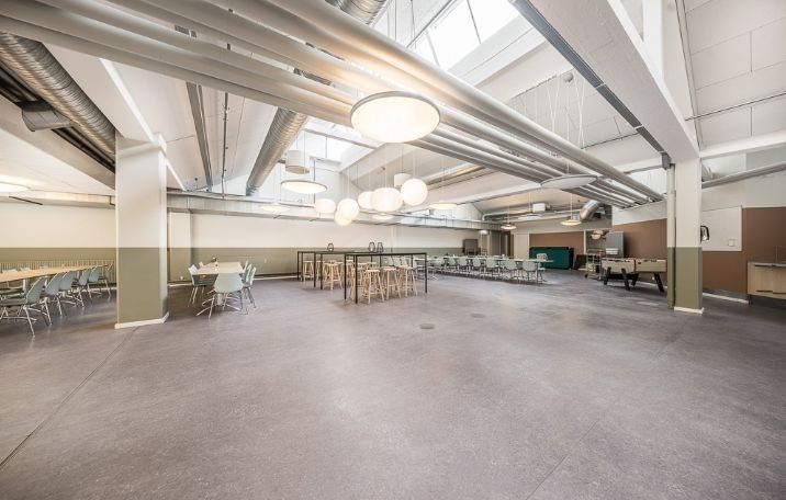 Store mødelokale med åben gulvplan og lyse loftslamper, lange runde borde og stole samt spilbord og sofaer