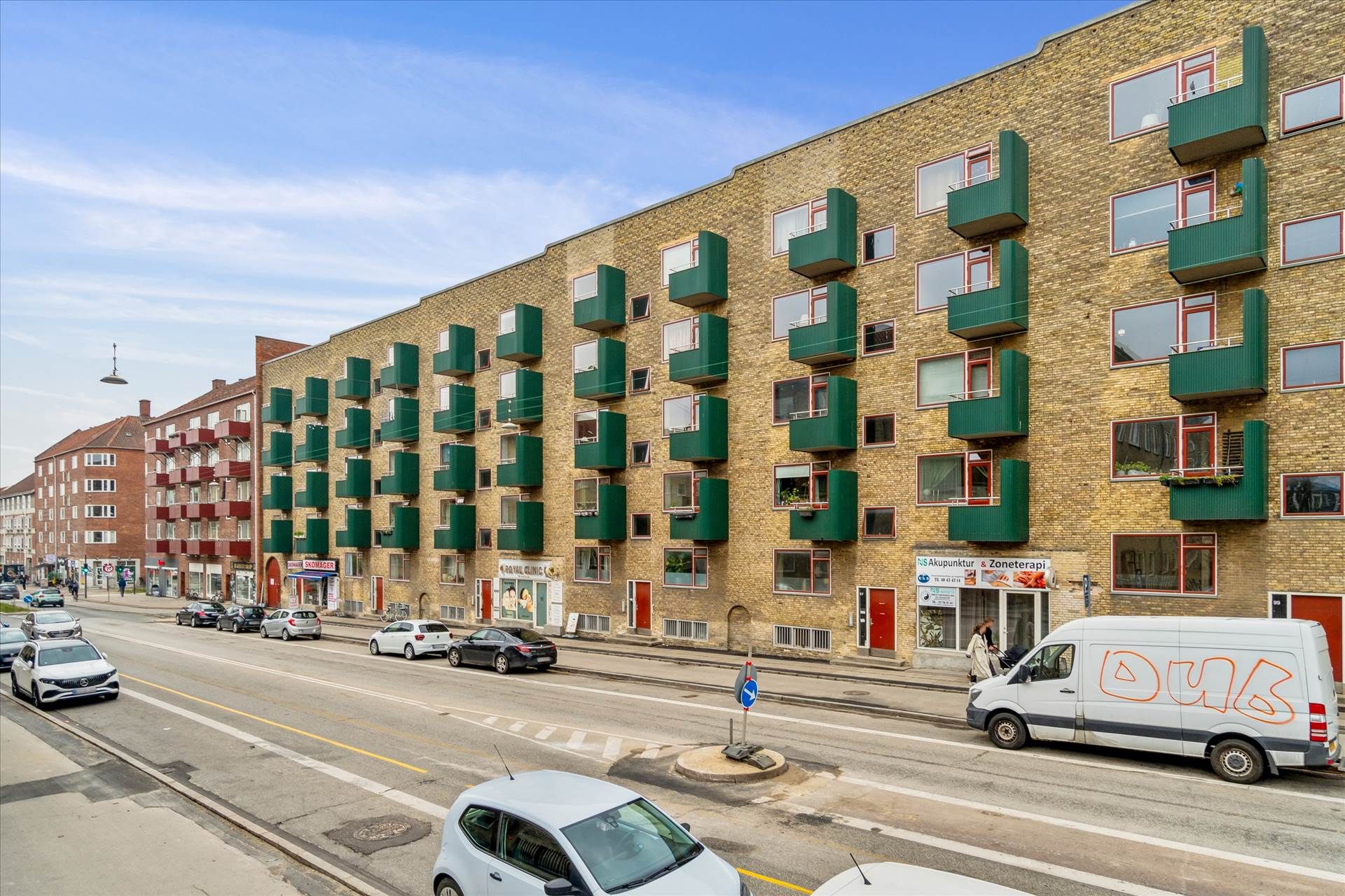 Lejlighedsbygning med grønne altaner og rød accent. Forretninger og biler på hjørnet. Rød og hvid bus og biler kører på gaden