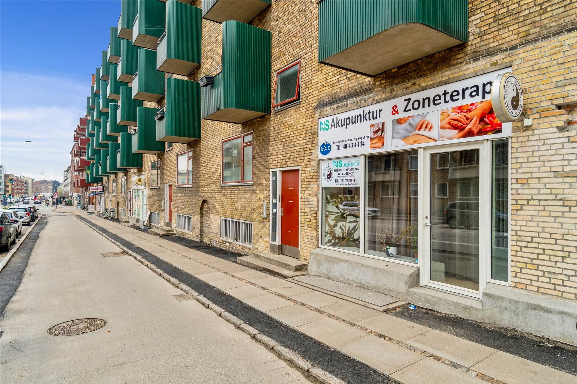 En tom gade med butikker og lejligheder, hvor den ene butik hedder Akupunktur & Zoneterapi med indgangsdør og vinduer med
