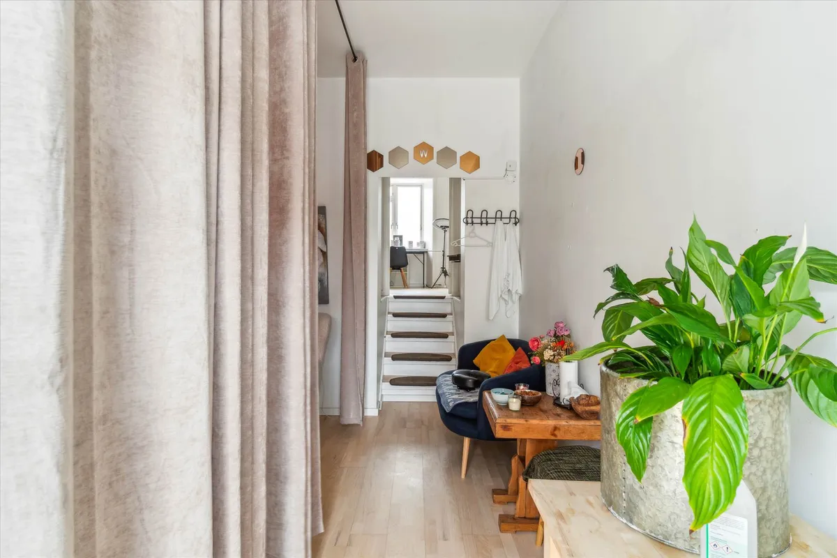 En indgangsparti med lyse gulv og vægge, møbler og indretning med planter og farverige puder samt en trappe.