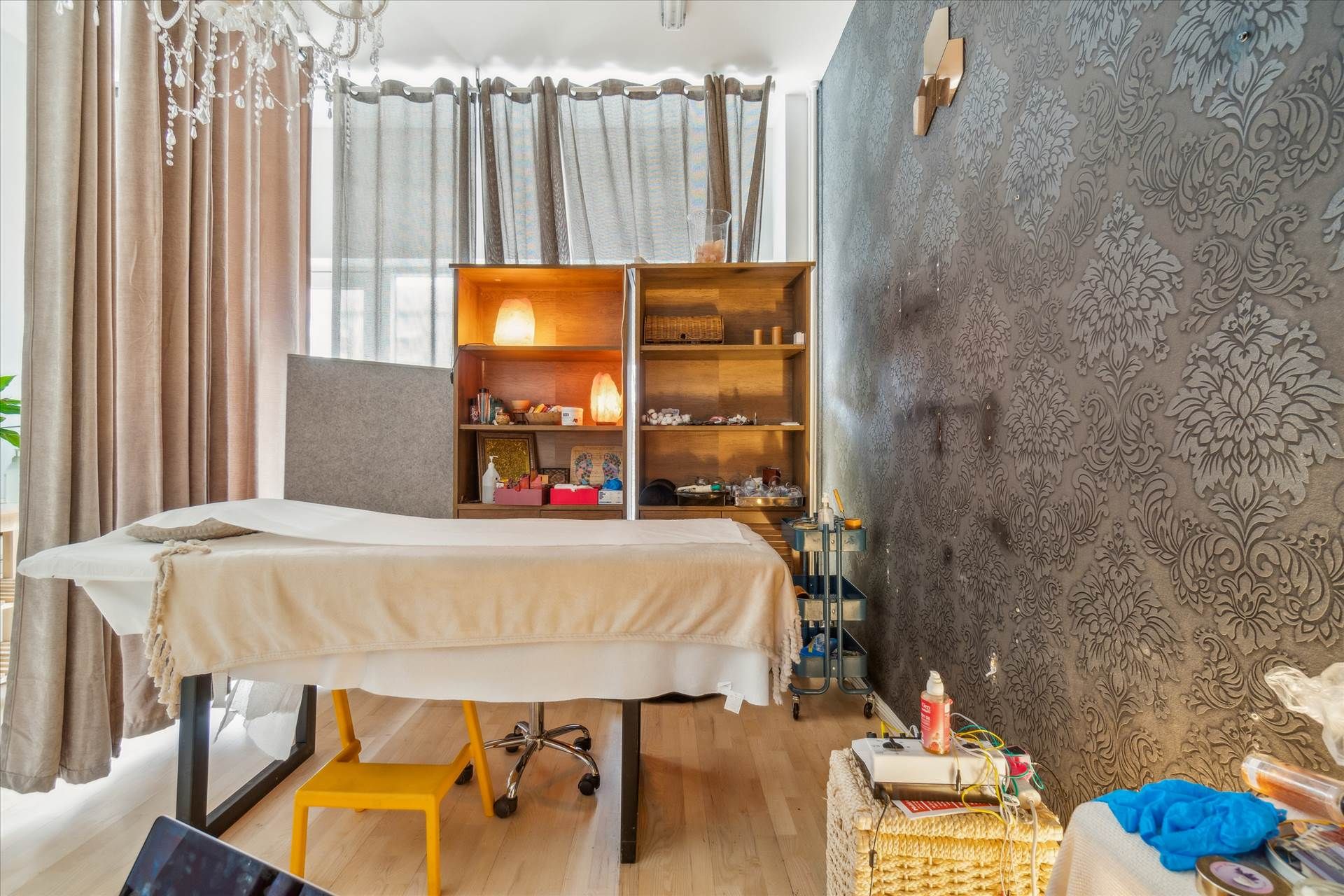 Massagebord og gul stol i lyseværelse med lysekroner og gardiner. Forskelligt udstyr og ting på hylder og væg.