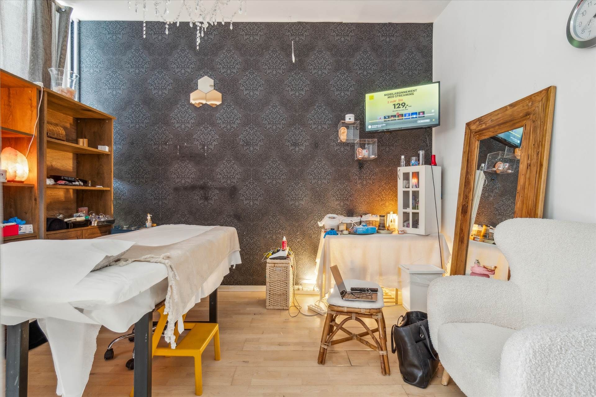 Massageværelse med massagebord og stol samt et skrivebord med bærbar computer og flere hylder med diverse ting.