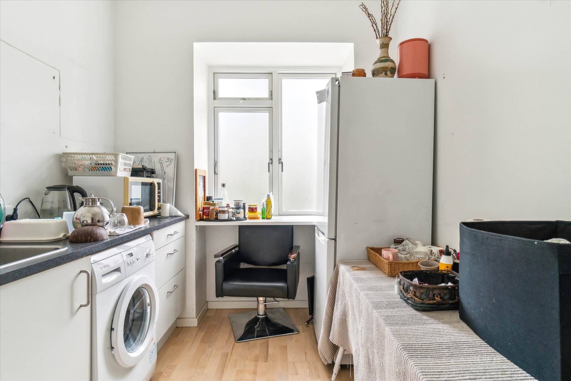 Køkken med hvidt køkkenskab, lysegrå gulv, hvid køleskab, stol, træbord og tørretumbler samt diverse husholdningsartikler på