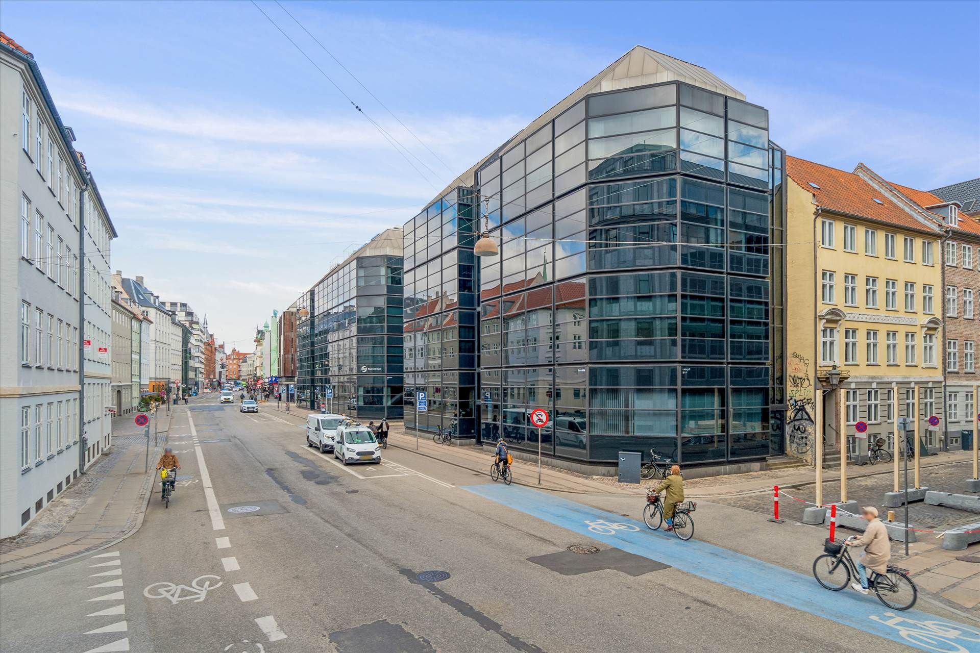 På et tomt vejafsnit kører folk på deres cykler i dagslys. Bygninger med glasvinduer og røde tag. En person cykler på en blå