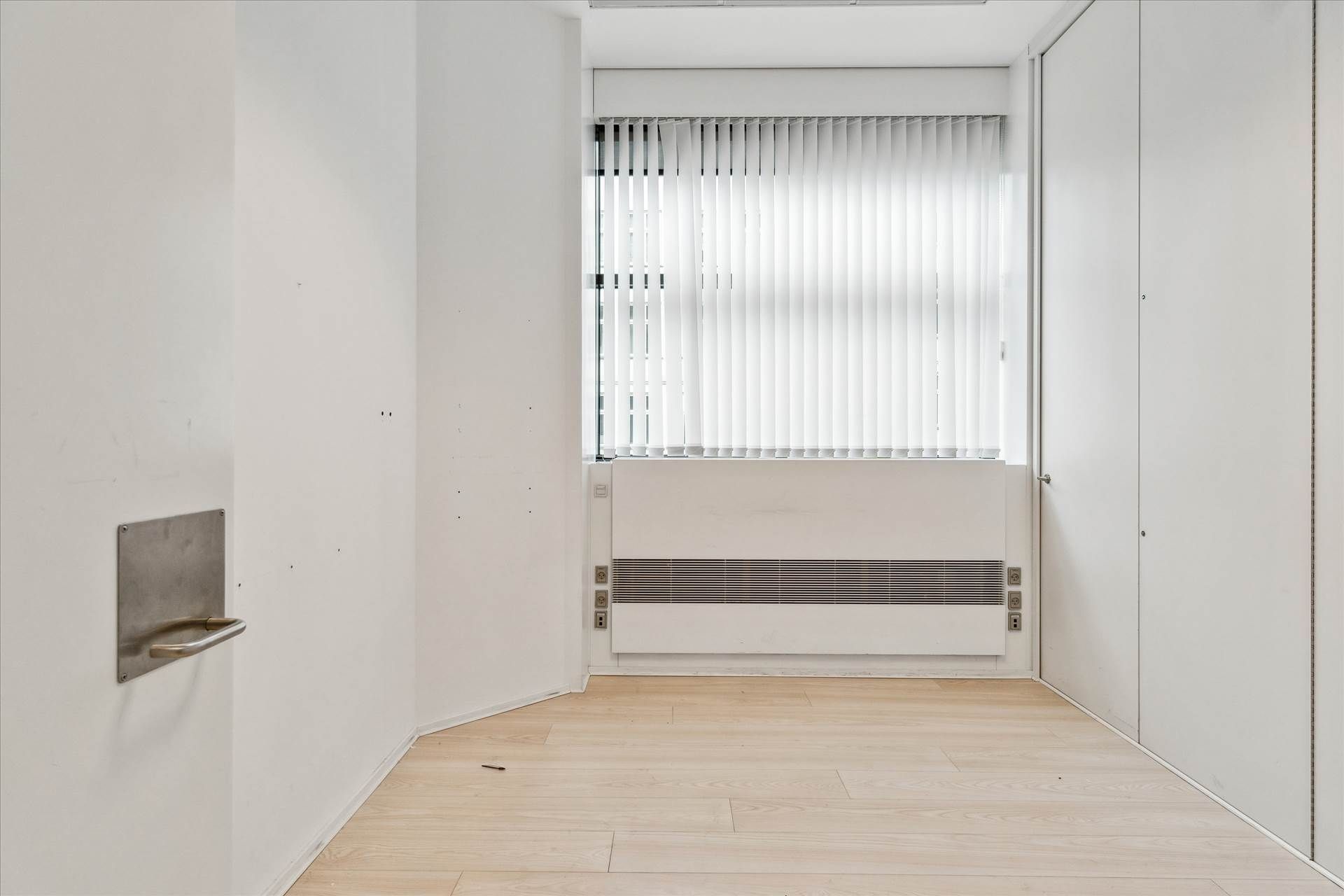 Lejlighed i stueetagen med møbleret køkken og lyse gulve, hvide vægge og hvide jalousier samt en stor, lysevinduesfløj.