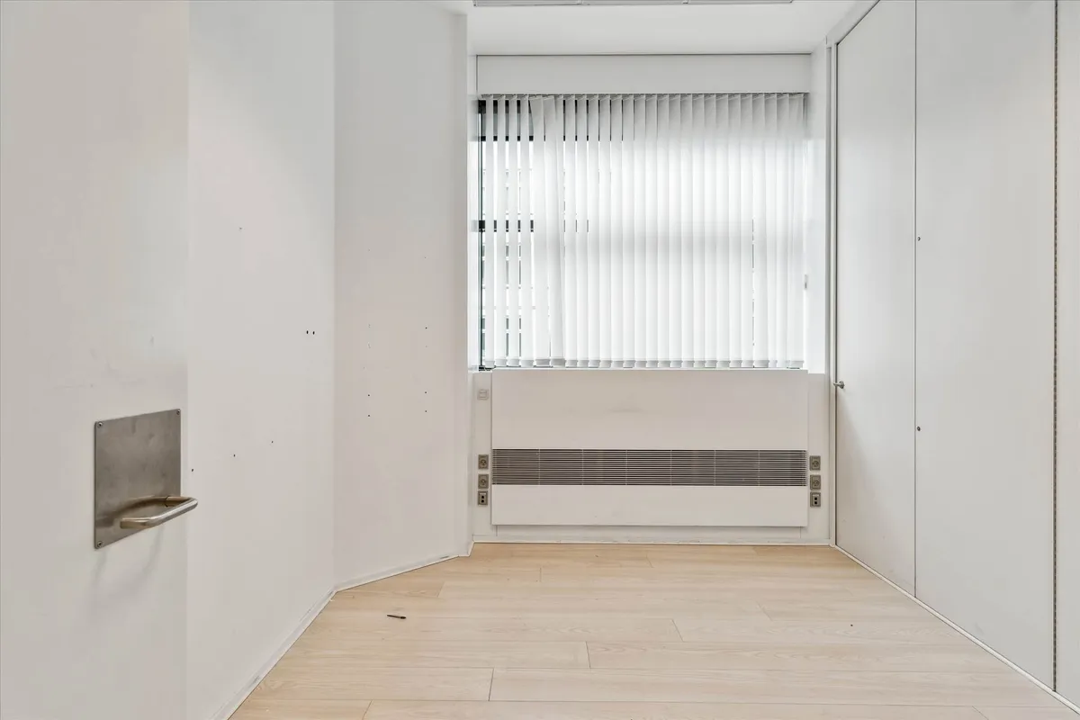 Lejlighed i stueetagen med møbleret køkken og lyse gulve, hvide vægge og hvide jalousier samt en stor, lysevinduesfløj.